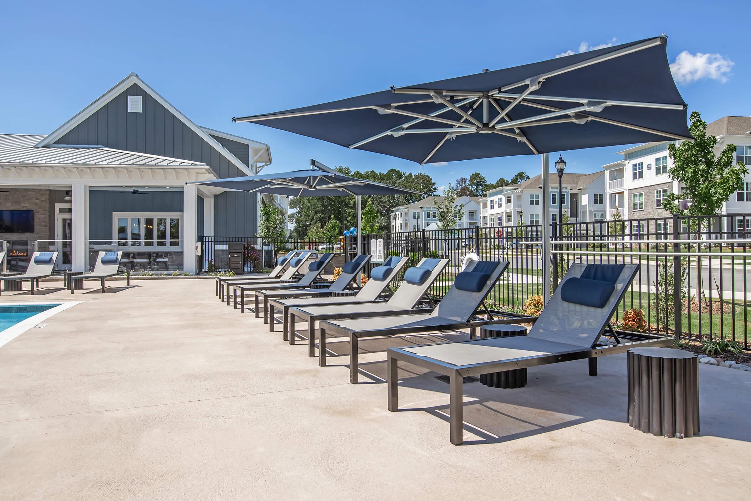 A modern outdoor pool area featuring several lounge chairs with blue cushions under large umbrellas. In the background, a stylish building can be seen alongside landscaped green areas and additional apartments. The scene is bright and inviting, perfect for relaxation.