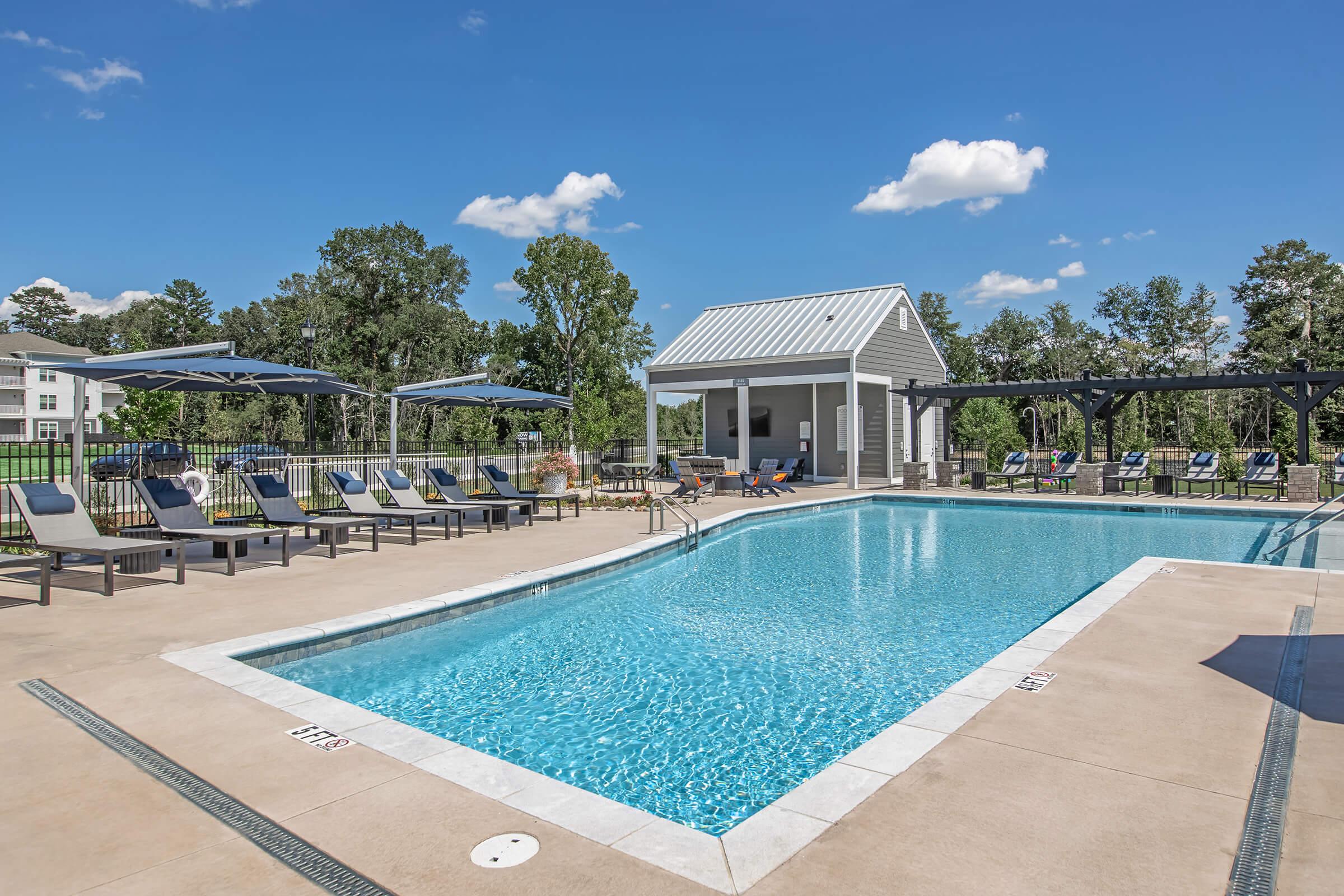 A bright outdoor pool area featuring a clear blue swimming pool surrounded by lounge chairs and shaded cabanas. A modern pool house is visible in the background, along with lush trees and a clear blue sky with a few clouds. The scene is inviting and designed for relaxation and leisure.