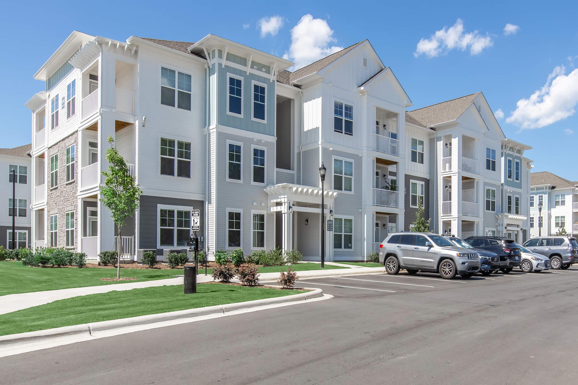A modern multi-story apartment building with white siding and gray accents, featuring balconies and large windows. The surrounding area includes well-maintained landscaping with trees and shrubs, and a parking lot with several vehicles. The sky is clear with a few clouds.