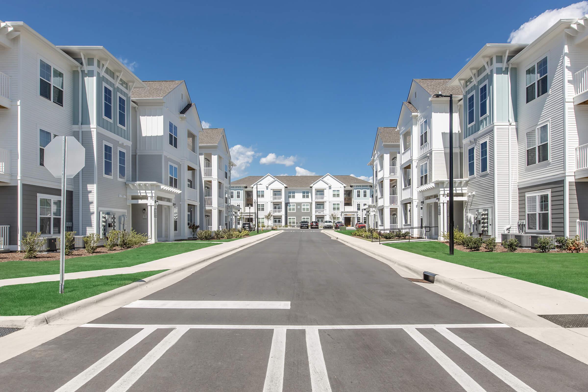 A wide view of a residential street lined with modern apartment buildings, featuring manicured lawns, clear blue skies, and a fresh, clean pavement. The scene includes crosswalk markings and street signs, creating a welcoming suburban atmosphere.