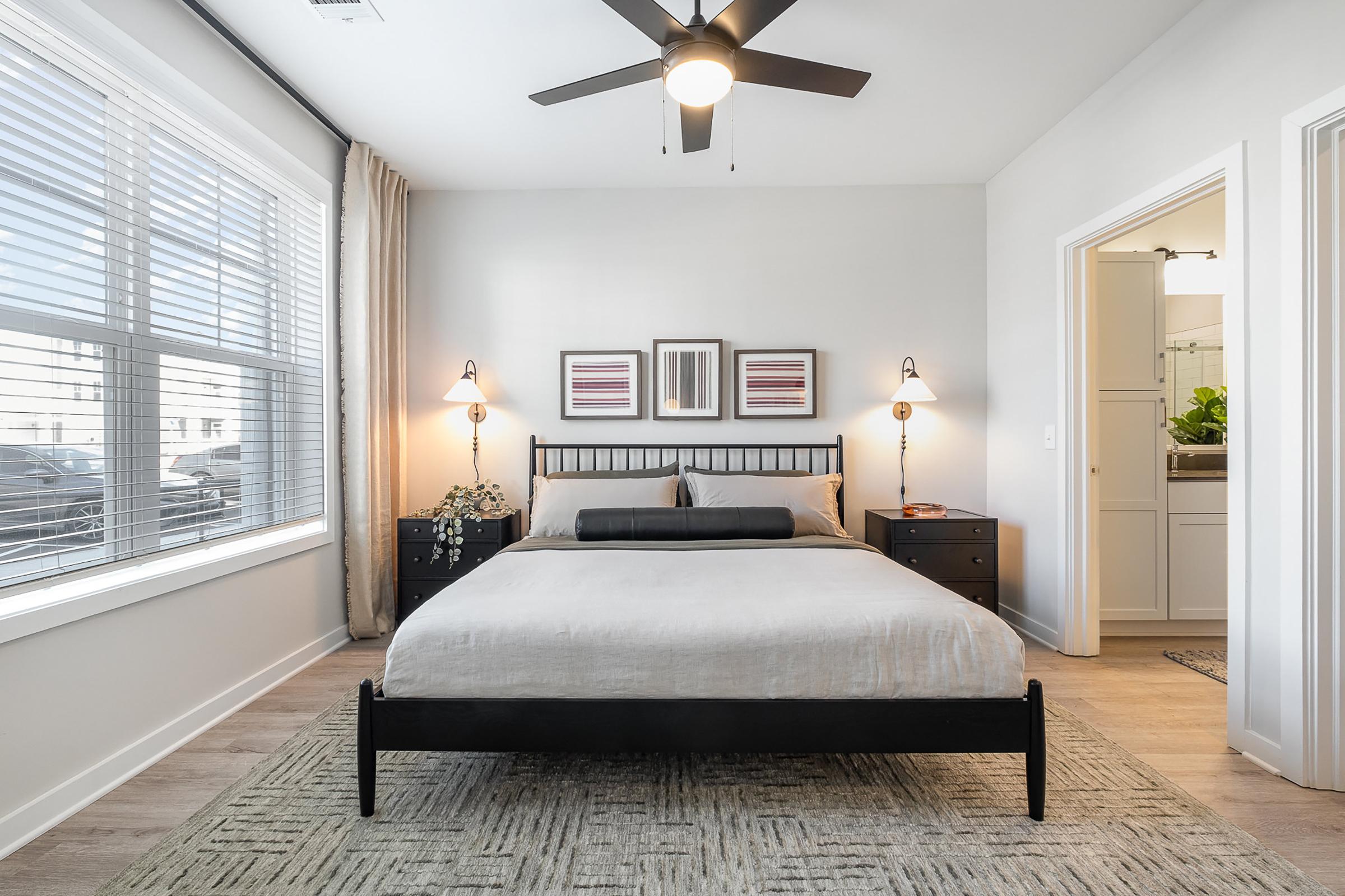 A modern bedroom featuring a queen-sized bed with light bedding against a neutral wall. There are two bedside tables with lamps, and artwork hanging above the bed. A large window allows natural light to fill the room, and a bathroom can be seen through an open door. The floor is covered with a textured rug.