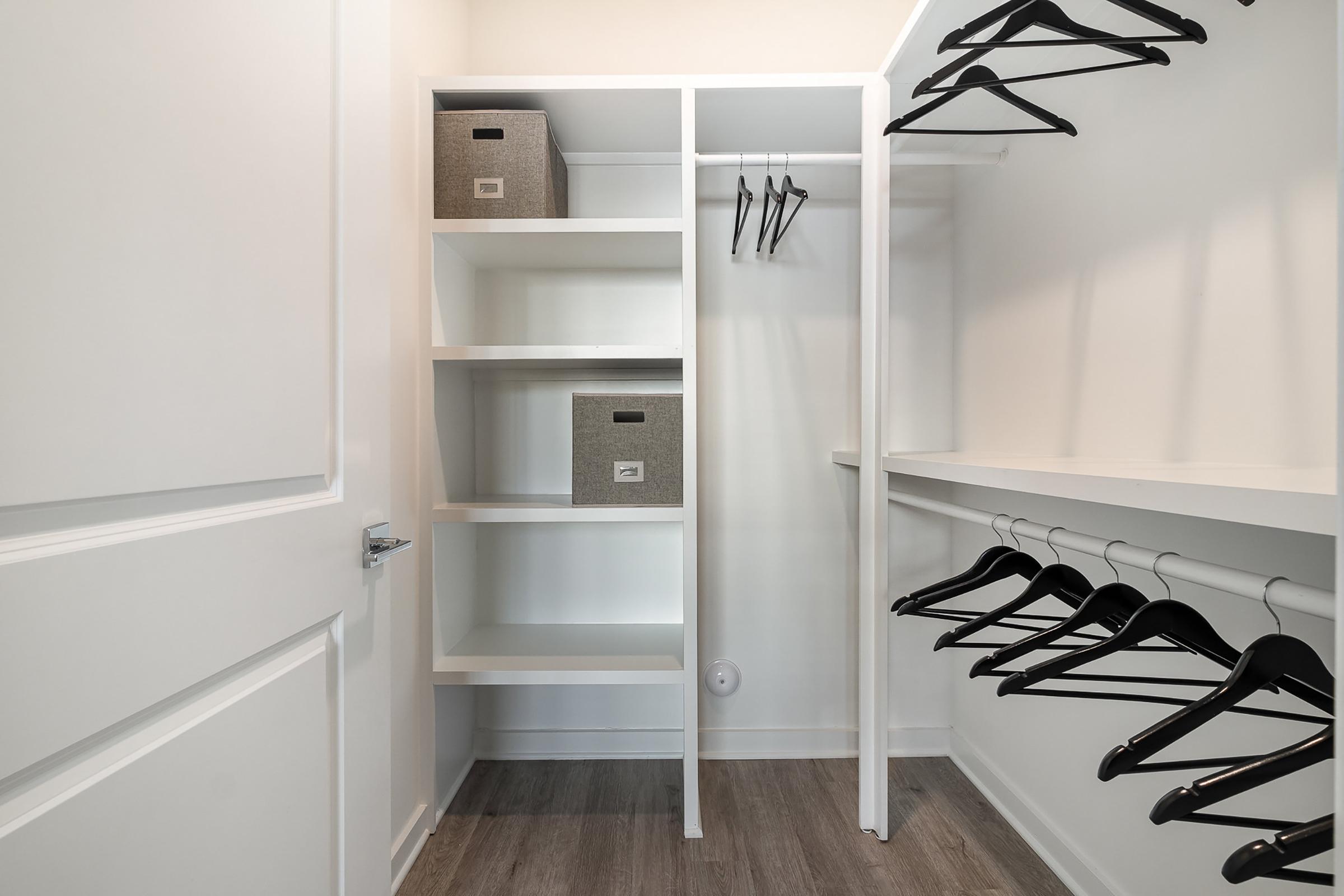 A well-organized closet featuring white shelving units with gray storage boxes and a smooth floor. Several black hangers are neatly arranged on a rod, with ample empty space suggesting it's ready for clothing or accessories. The walls are painted in a light color, creating a bright and tidy atmosphere.