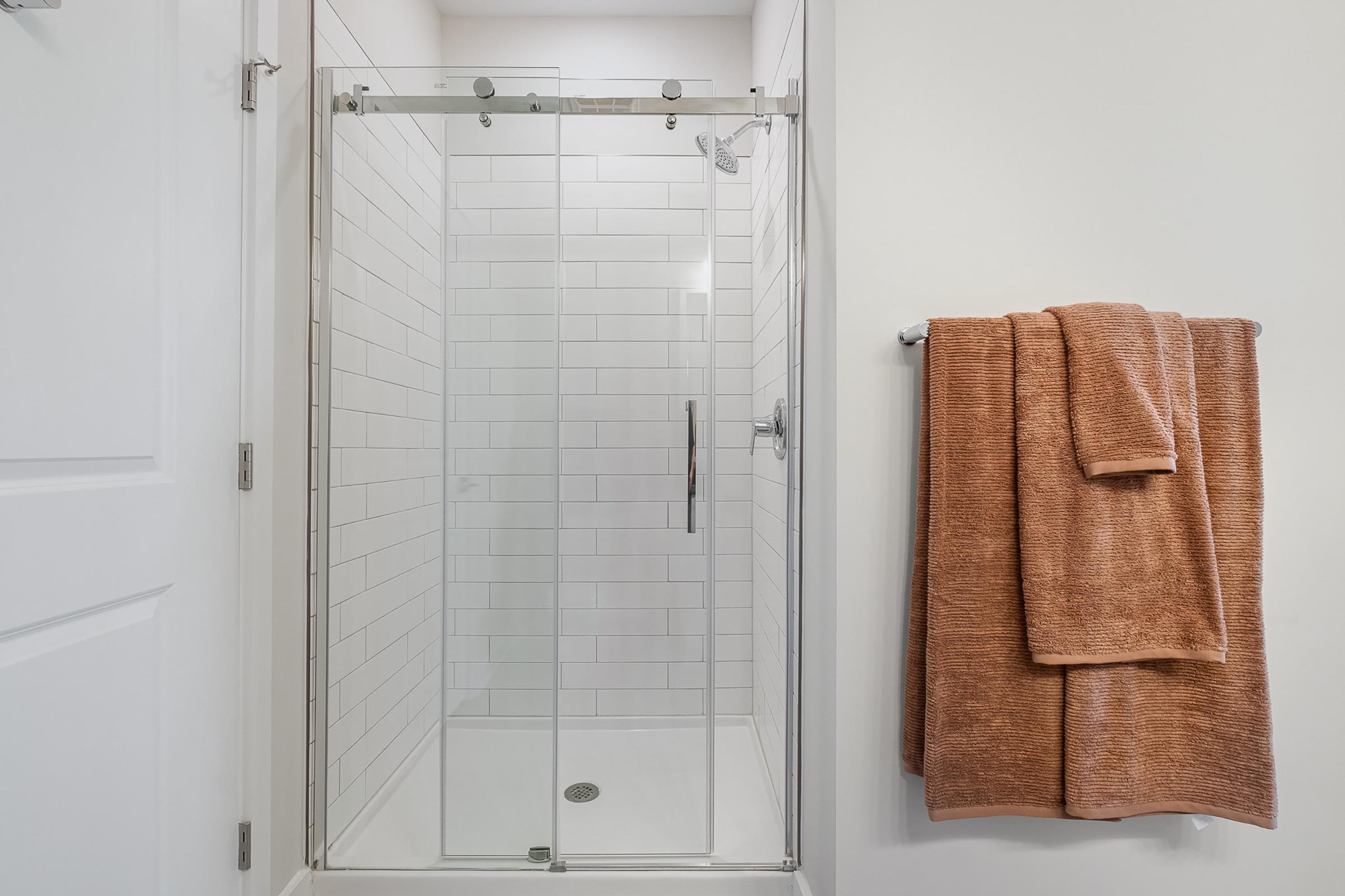 A modern bathroom featuring a glass shower enclosure with sliding doors, white tiled walls, and a showerhead. A neatly hung orange towel is on a towel bar next to the shower, and the overall color scheme is bright and clean.