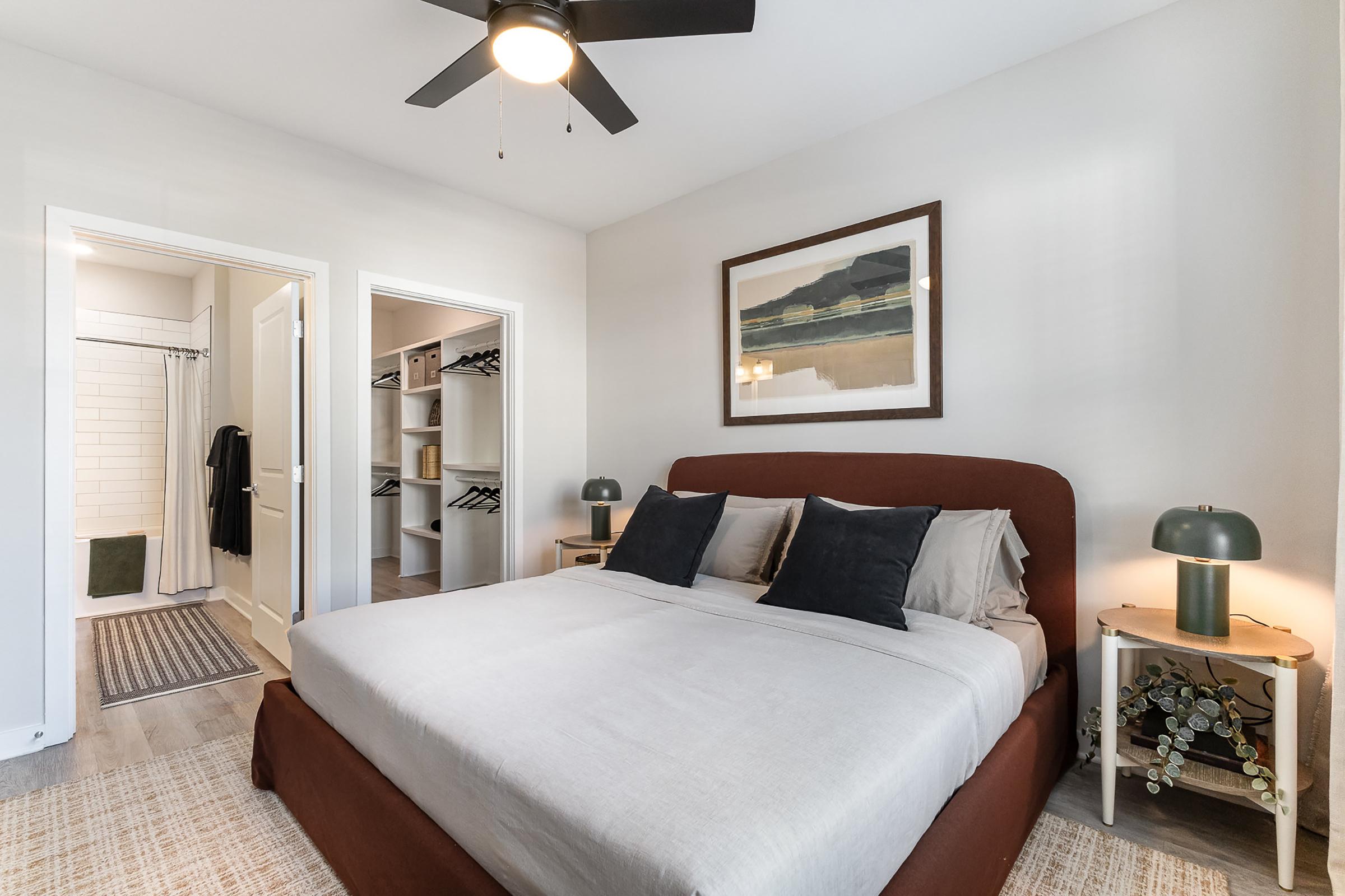 Cozy bedroom featuring a bed with beige linens and dark pillows, two bedside lamps, and framed artwork on the wall. A hallway leads to a walk-in closet and a bathroom with a shower, all accented by neutral tones and a ceiling fan. The flooring is light wood with a textured area rug.