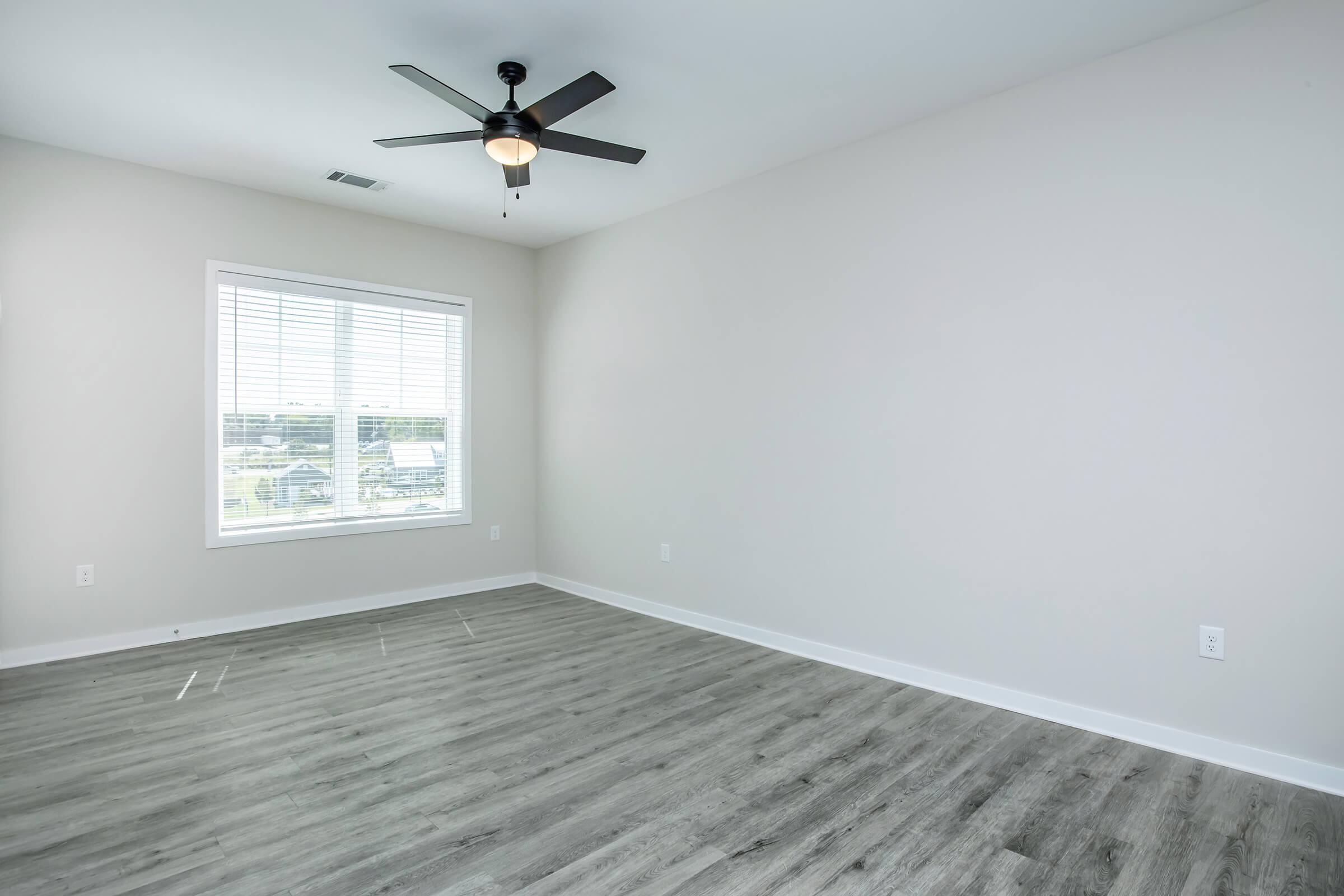 Empty room with light gray walls and a ceiling fan. There is a large window with blinds allowing natural light to enter, and the floor is covered with light wood-like flooring. The space appears modern and clean, suitable for various uses.