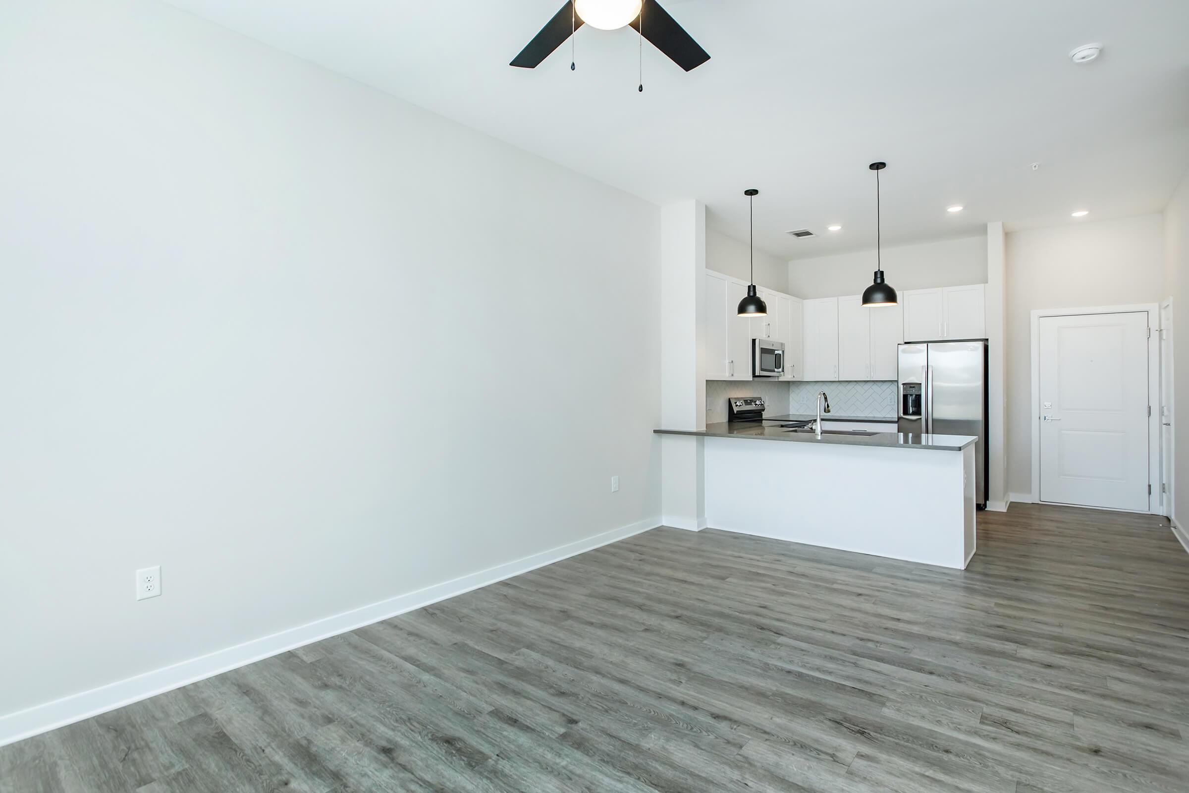 A spacious, modern kitchen and living area with light-colored walls and wood-style flooring. The kitchen features stainless steel appliances, white cabinetry, and a breakfast bar. Two black pendant lights hang above the bar, and a ceiling fan is visible. The room is bright with ample natural light.
