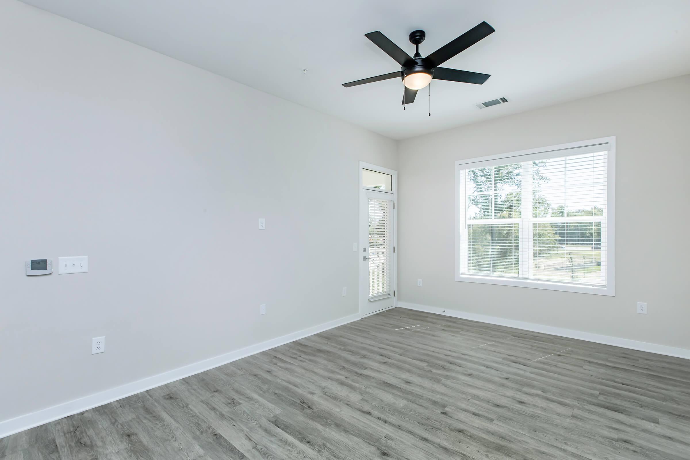 Empty room featuring light-colored walls, a ceiling fan, and large window with blinds. The floor is covered in light hardwood, and there is a door leading outside. Natural light illuminates the space, creating a bright and airy atmosphere.