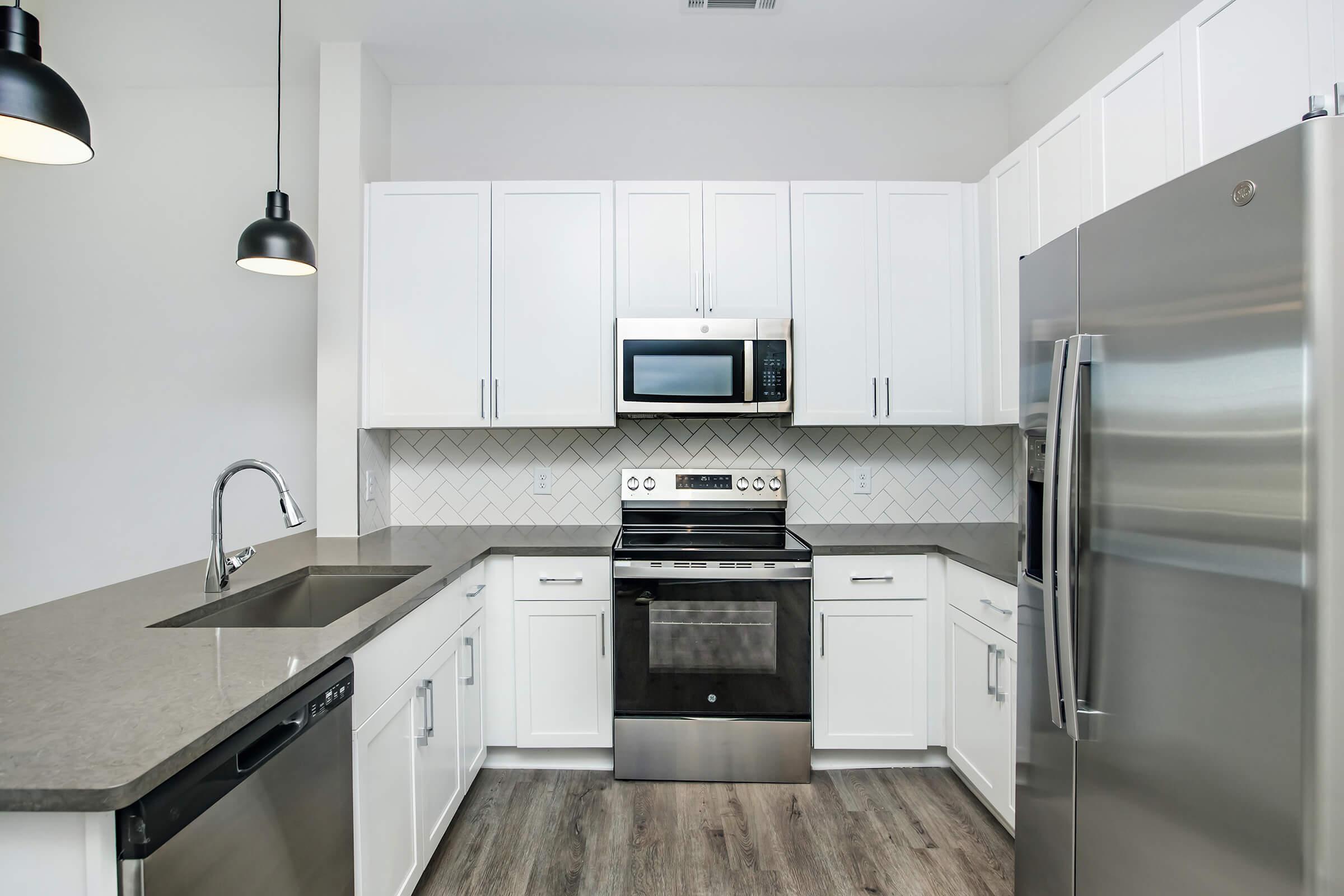 Modern kitchen featuring sleek white cabinetry, a stainless steel refrigerator, oven, and microwave. The countertop is gray with a herringbone-patterned backsplash. A stainless steel sink is integrated into the counter, and pendant lights hang above, illuminating the space. The floor is light wood.