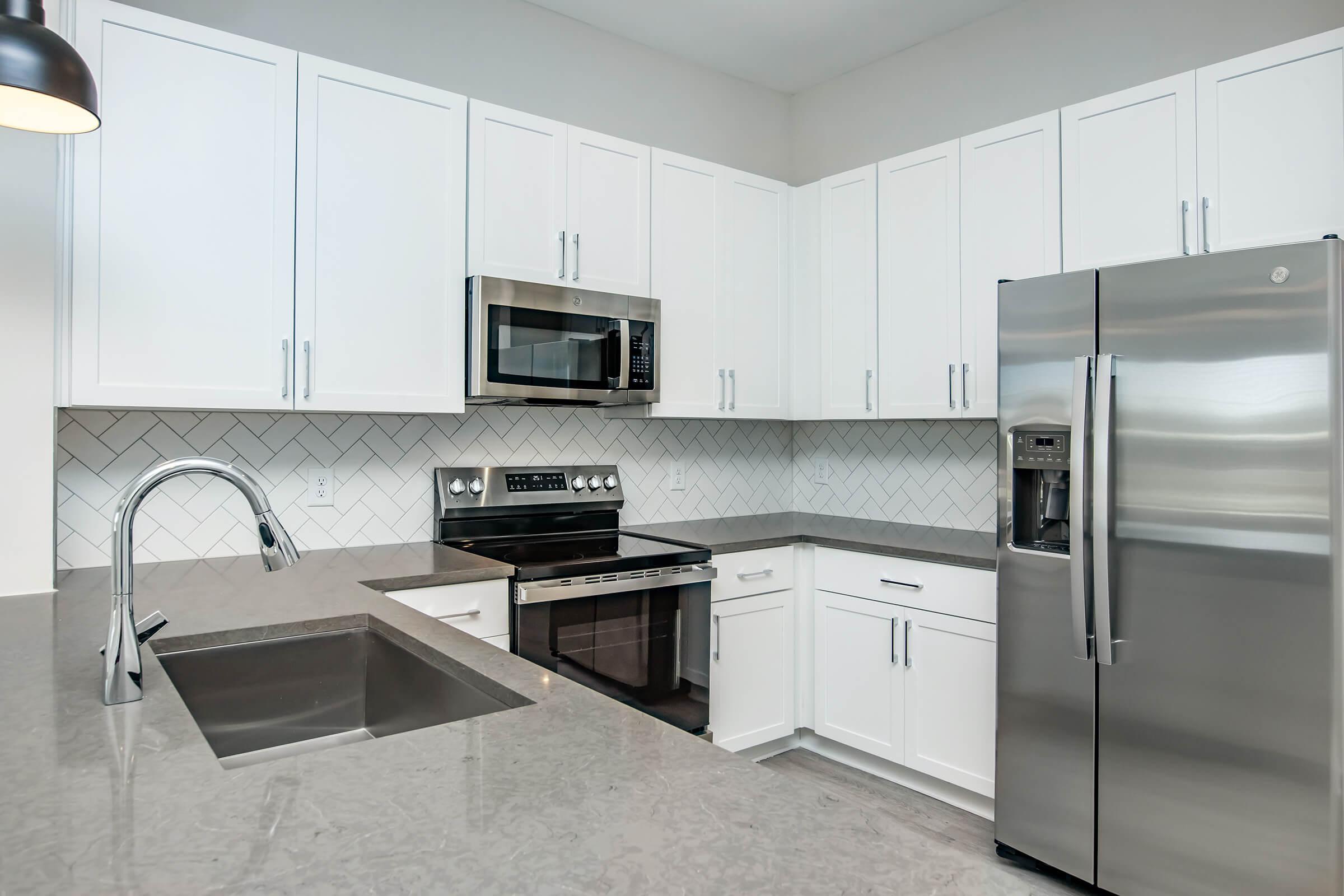 Modern kitchen with white cabinetry, stainless steel appliances, and a gray countertop. The space features a built-in microwave, an oven with stovetop, a refrigerator with ice and water dispenser, and a double-basin sink with a sleek faucet. The backsplash is designed with herringbone-patterned tiles.