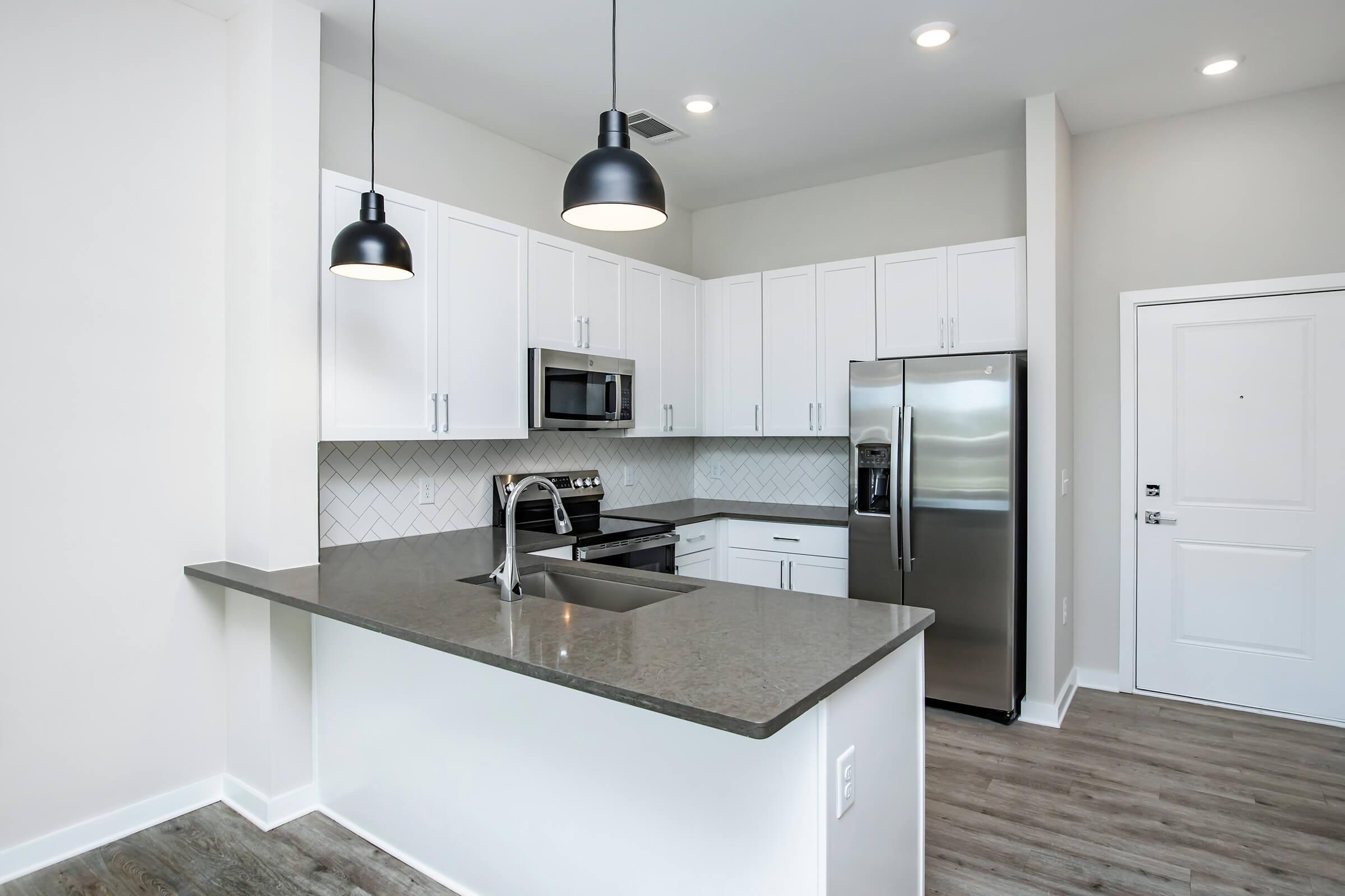 Modern kitchen featuring white cabinetry, stainless steel appliances, a large granite countertop with seating, and pendant lighting. The walls are painted in a light color, and there is a herringbone-patterned backsplash. A door is visible, likely leading to another room or exterior.