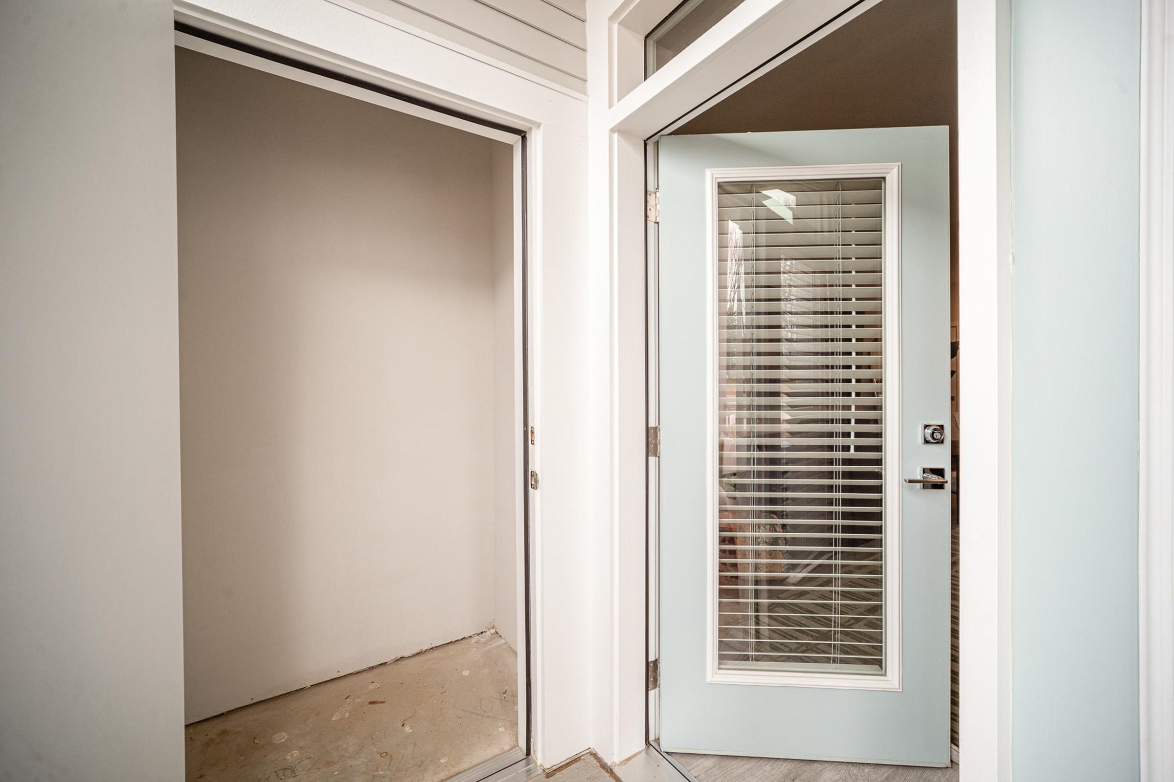 A contemporary entryway featuring a light blue door with glass panels, leading to a small room on the left with empty walls and visible flooring. The entrance has clean lines and modern design elements, highlighting a minimalist aesthetic. Natural light may be filtered through the door's glass.