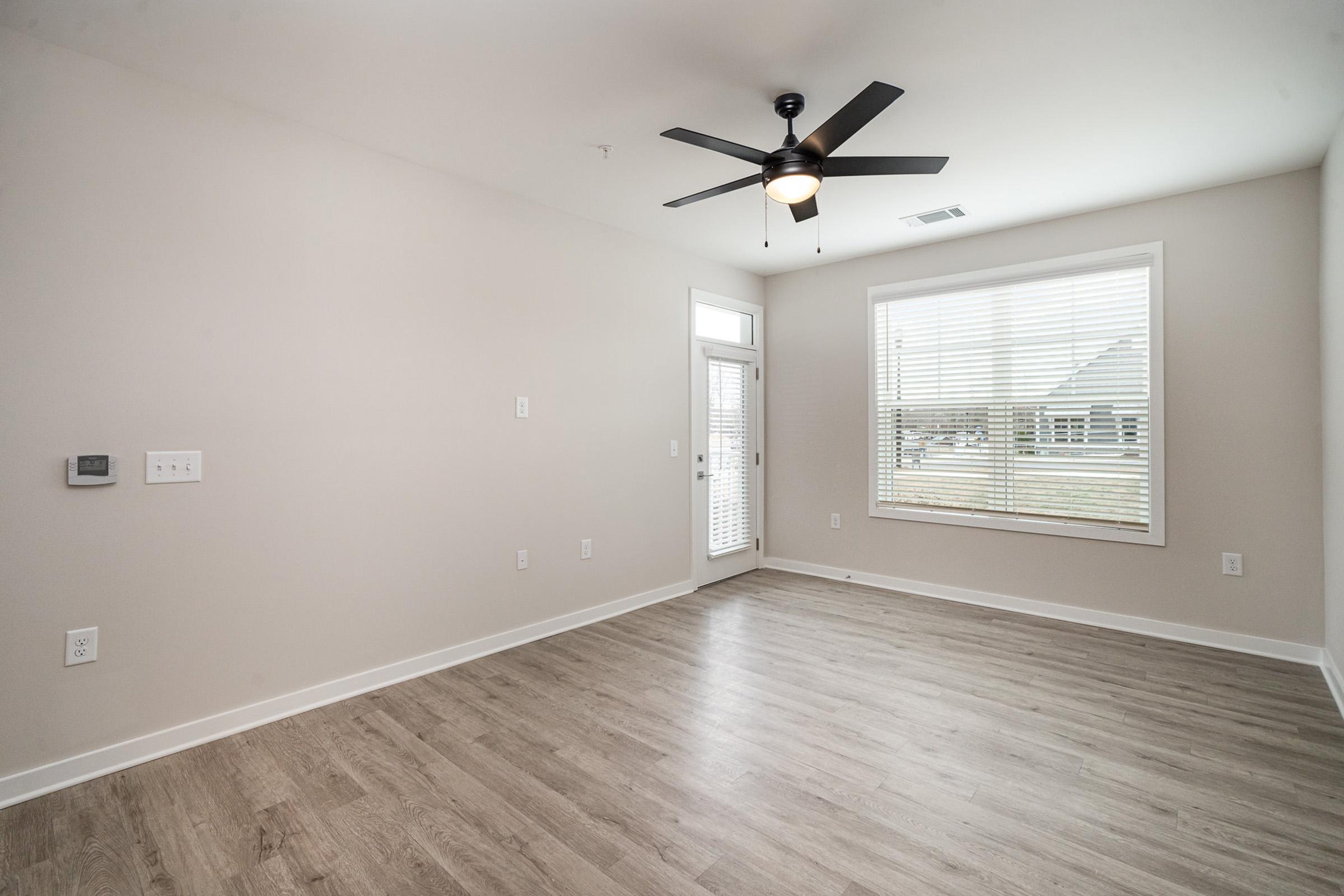 A spacious, empty room featuring light-colored walls, a ceiling fan, and large windows with blinds. The floor is hardwood, and there is a door leading outside. The overall atmosphere is bright and airy, suitable for various interior designs.