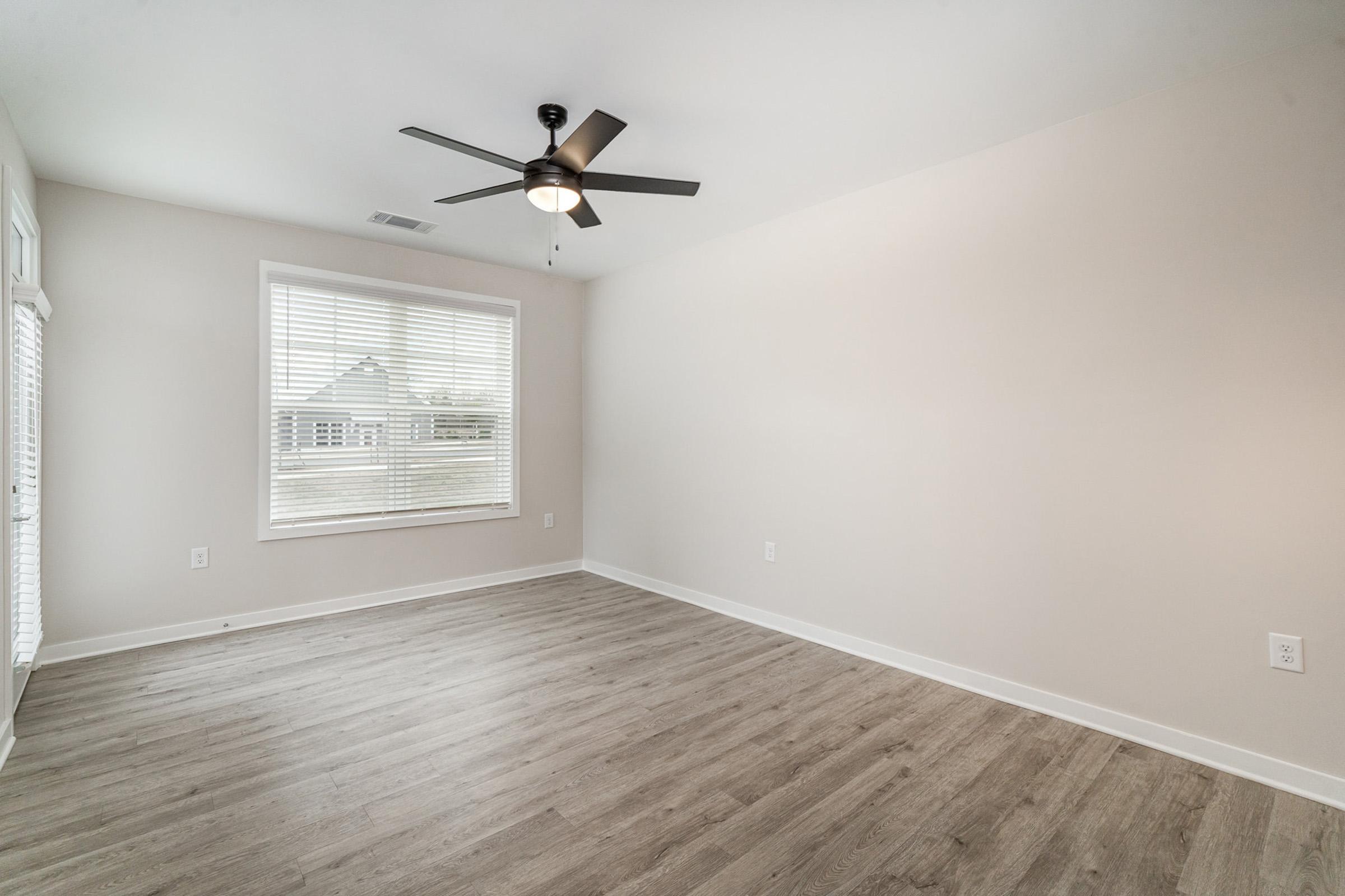 A bright, minimalistic room with light-colored walls and hardwood flooring. A ceiling fan is mounted in the center, and a large window with blinds allows natural light to enter. The space appears empty, suggesting it could be used as a living room or bedroom.