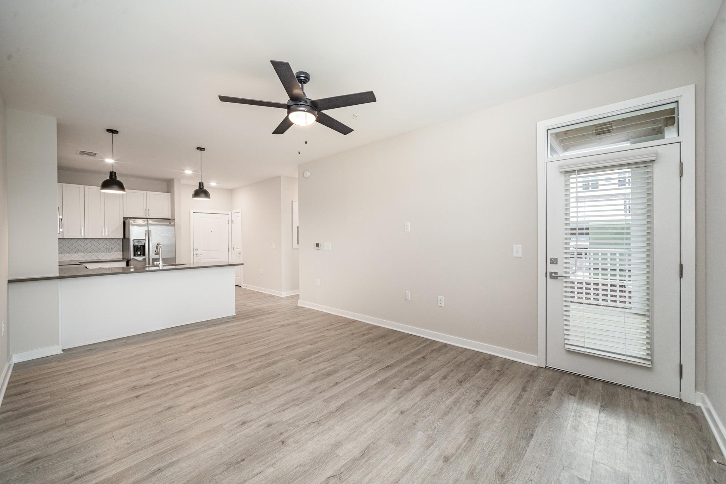 A modern, spacious living area featuring light wood flooring, a ceiling fan, and large windows. The space includes a doorway leading to the outside and a kitchen area with sleek cabinetry and stainless steel appliances in the background.