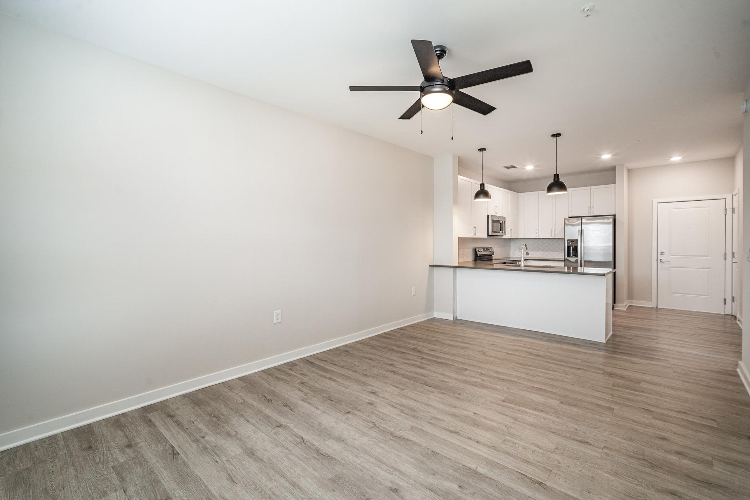 Modern open-concept living space featuring a kitchen with a breakfast bar, stainless steel appliances, and pendant lighting. The room has a ceiling fan, light hardwood flooring, and neutral-colored walls, creating an airy atmosphere. The entrance to the adjacent room is visible on the right.