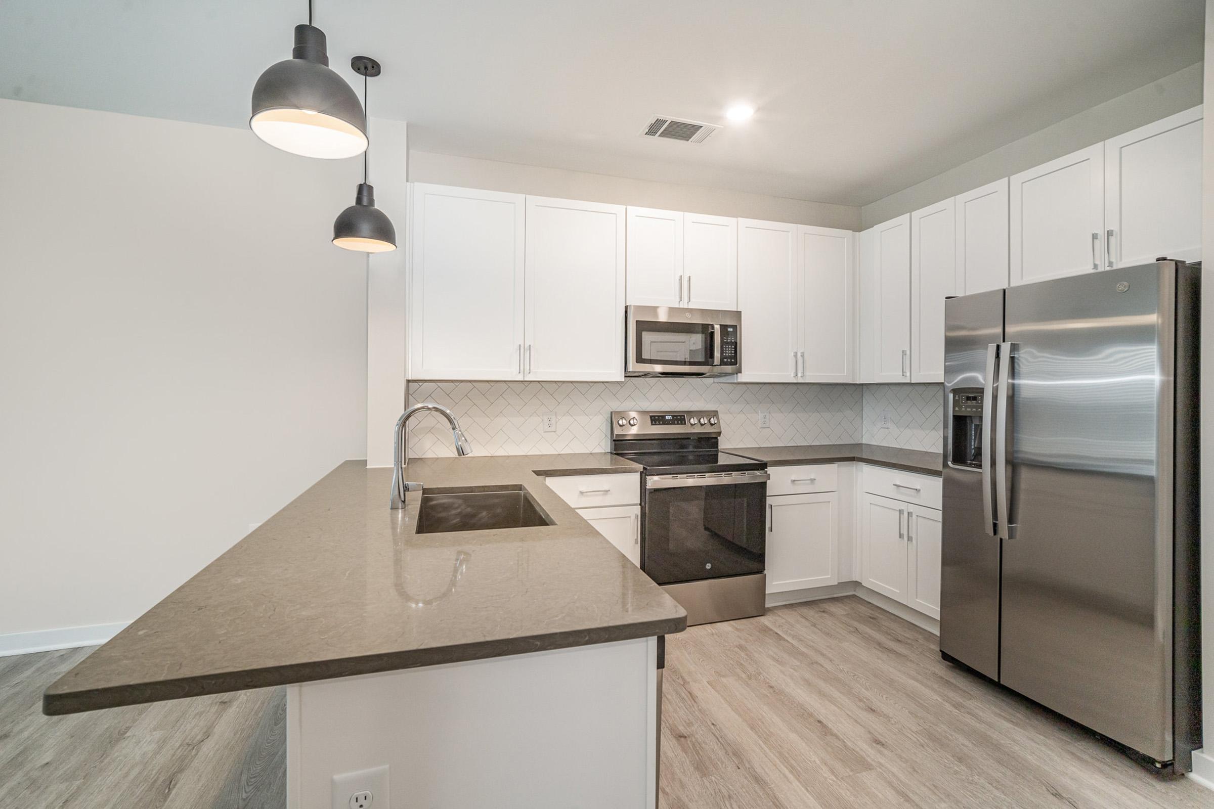 Modern kitchen featuring white cabinets, a stainless steel refrigerator, and an oven with a microwave above it. The countertop is dark, complemented by stylish pendant lighting above the island. The backsplash has a herringbone pattern, and the floor is light wood, creating a bright and contemporary atmosphere.