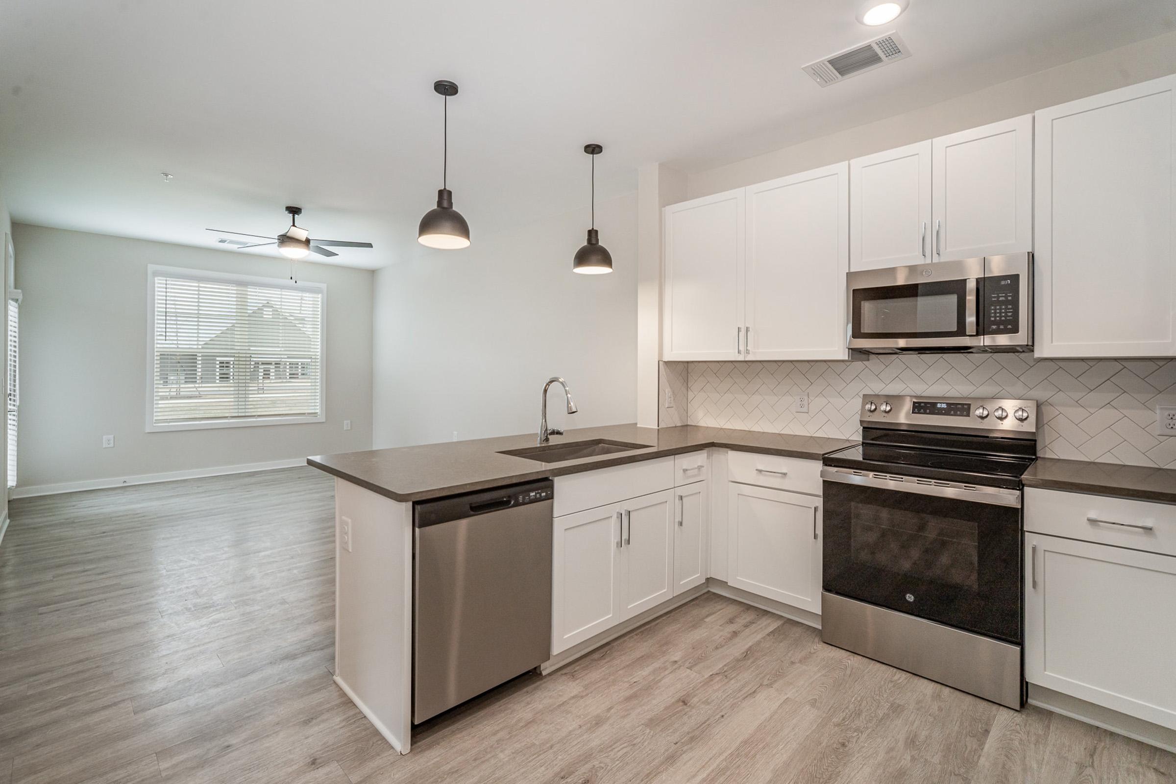 Modern kitchen with white cabinets, a stainless steel microwave and stove, a dishwasher, and a dark counter. Pendant lights hang above the counter. The open-concept space leads to a living area with a ceiling fan and large windows, offering natural light. The floor features light-colored wood laminate.