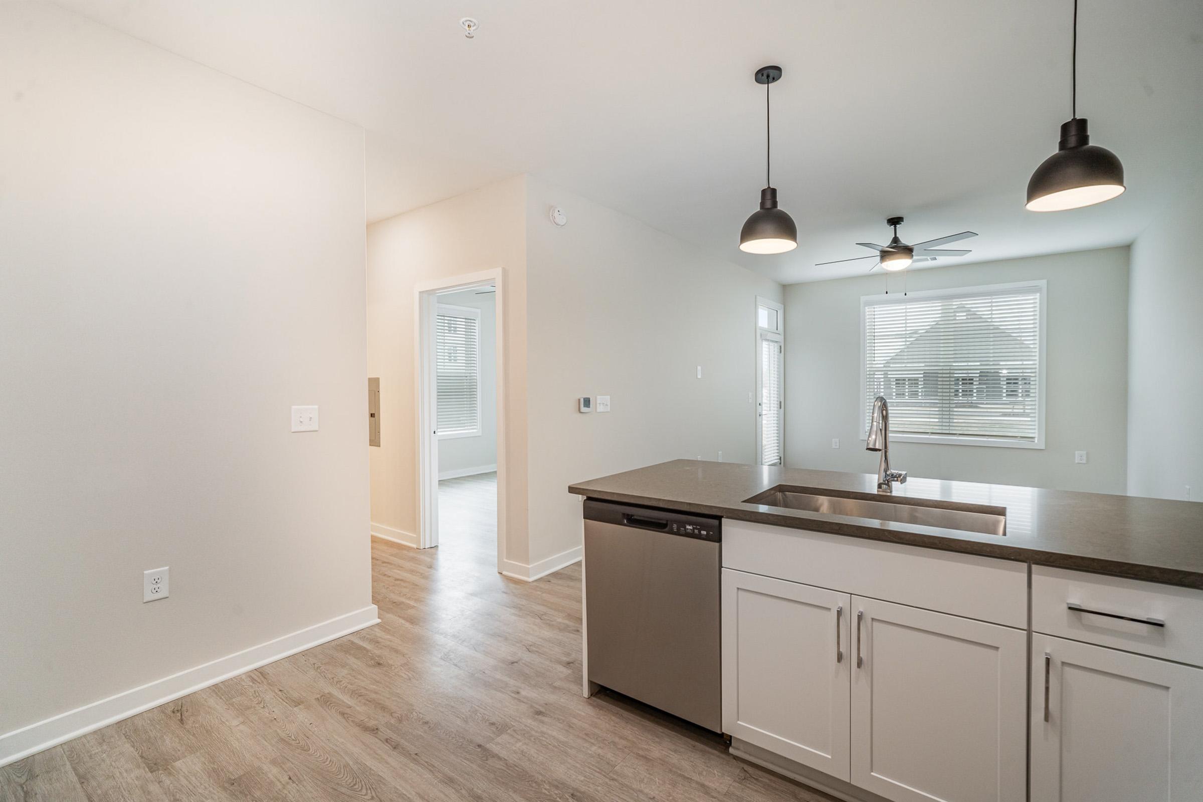 Modern kitchen interior featuring white cabinetry, a stainless steel dishwasher, and a large island with a dark countertop. There are pendant lights above the island, and the space is brightly lit with natural light coming from the nearby window. The room has a neutral color palette and wooden flooring.