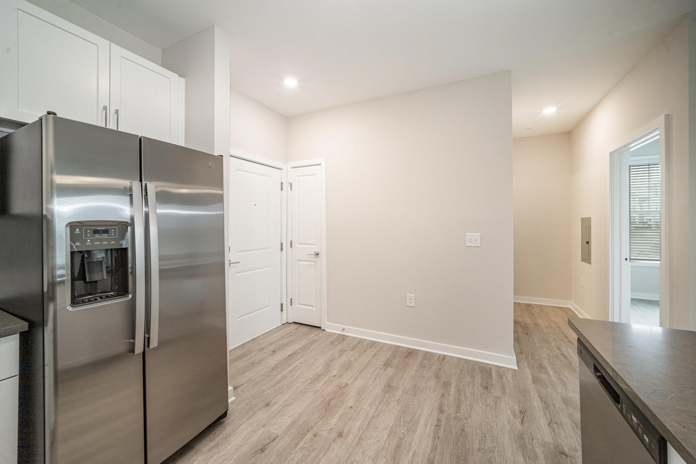 Modern kitchen showcasing stainless steel appliances, including a large refrigerator and dishwasher. The space features light-colored walls, wood-style flooring, and a doorway leading to another room. Bright lighting enhances the open and inviting atmosphere.