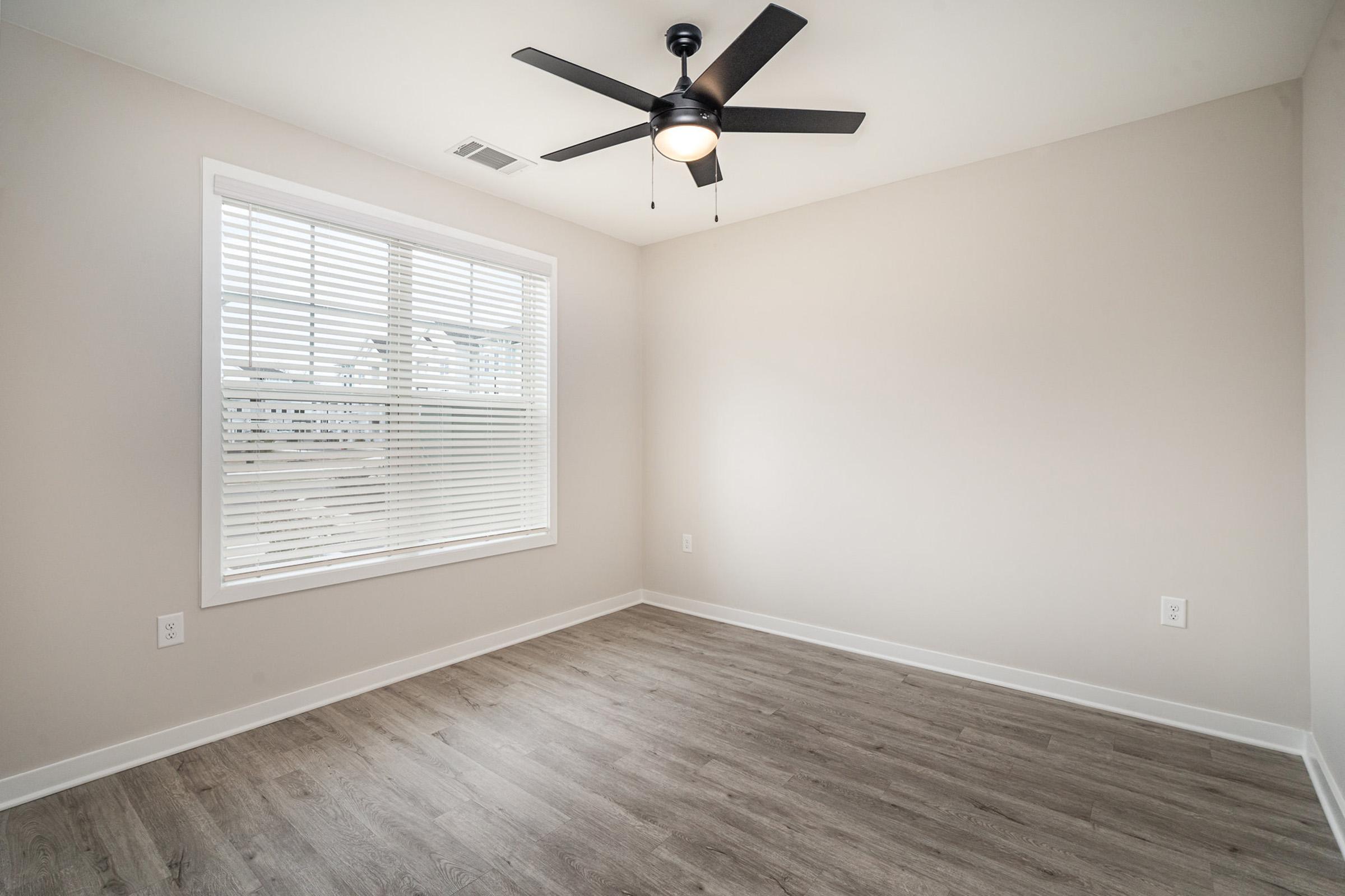 A spacious room with light beige walls and a wood-like floor. There is a ceiling fan with a light fixture, and a large window with white blinds allowing natural light to enter. The room appears empty, creating a clean and minimalistic atmosphere.