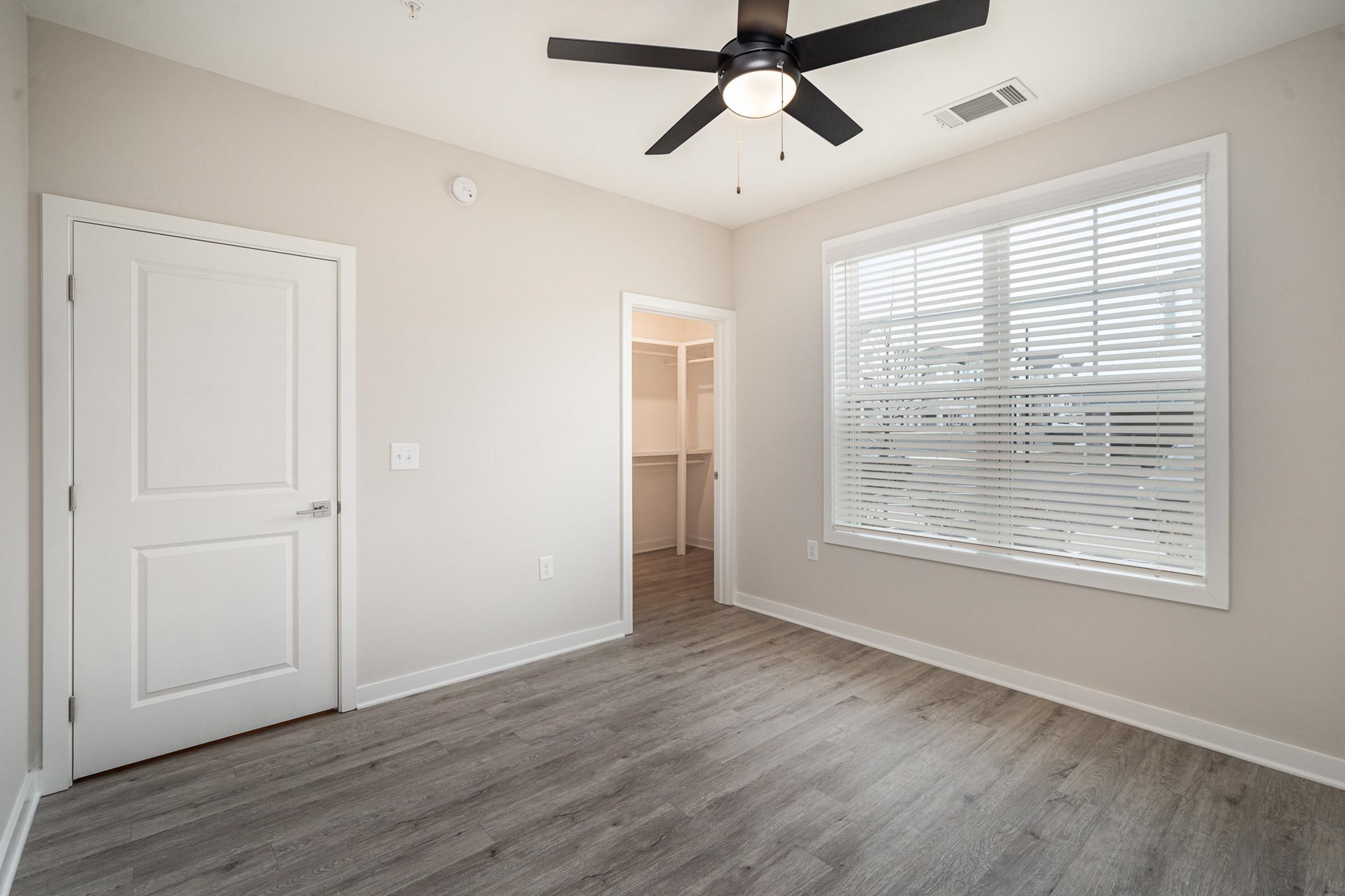 A well-lit, empty room featuring light-colored walls, a ceiling fan, and wood-like flooring. There is a closed door on the left, a large window with blinds allowing natural light, and a doorway leading to a closet. The space is clean and modern, ideal for personalizing.