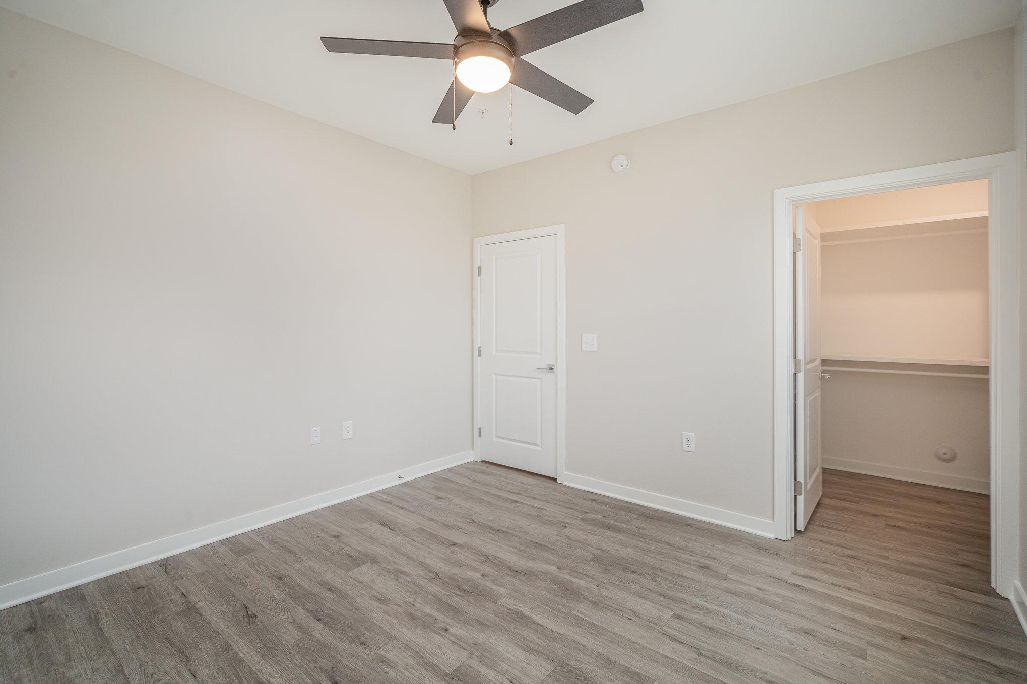 An empty room featuring light-colored walls, a ceiling fan with a light fixture, and light wooden flooring. There is a closed door on the left and an open closet with white shelving on the right. The space is well-lit and minimalistic, ideal for various uses.