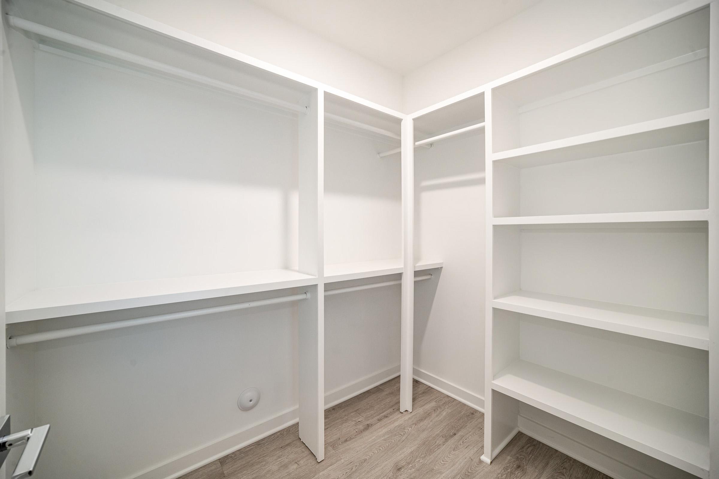 Empty walk-in closet featuring white shelving and rods for hanging clothes, with light-colored laminate flooring. The closet is well-lit and organized, with ample space for storage.