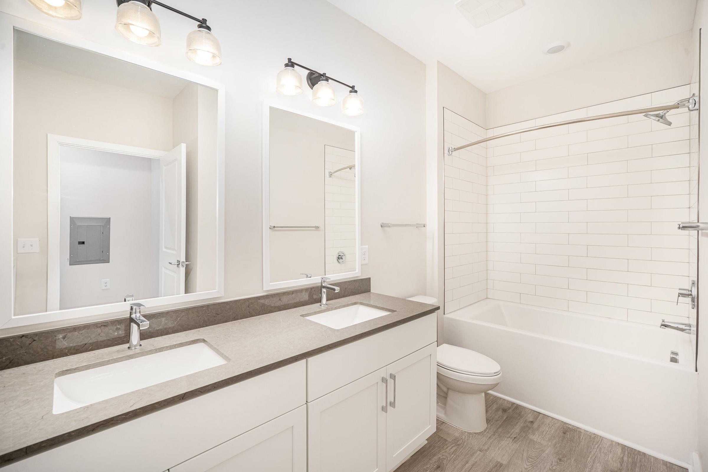 A modern bathroom featuring dual sinks with faucets on a dark countertop, two rectangular mirrors above the sinks, a white bathtub with a glass shower door, and light-colored tile walls. The space is bright and minimalistic, with neutral tones and good lighting.