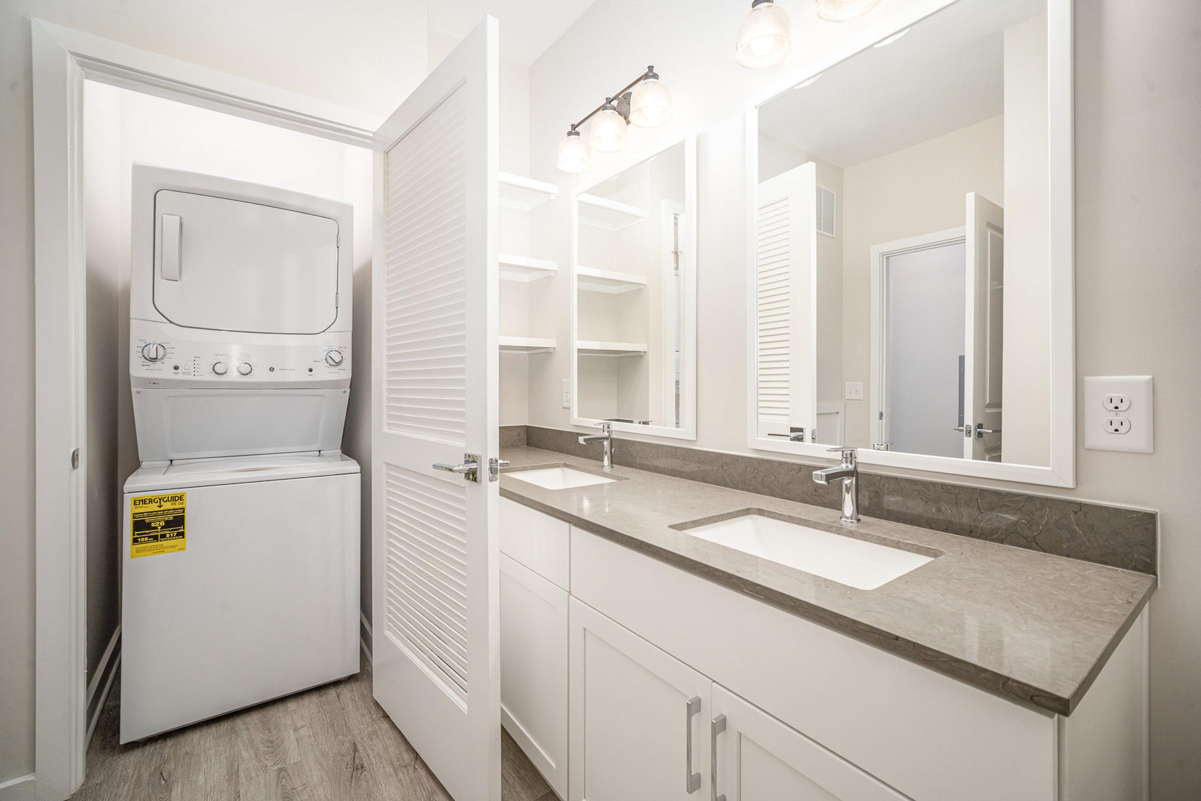 A modern bathroom featuring a double sink vanity with sleek fixtures, a mirror, and a stacked washer and dryer unit in a small alcove. The space has light-colored walls and wooden flooring, creating a bright and contemporary atmosphere. Adequate storage shelves are also visible.