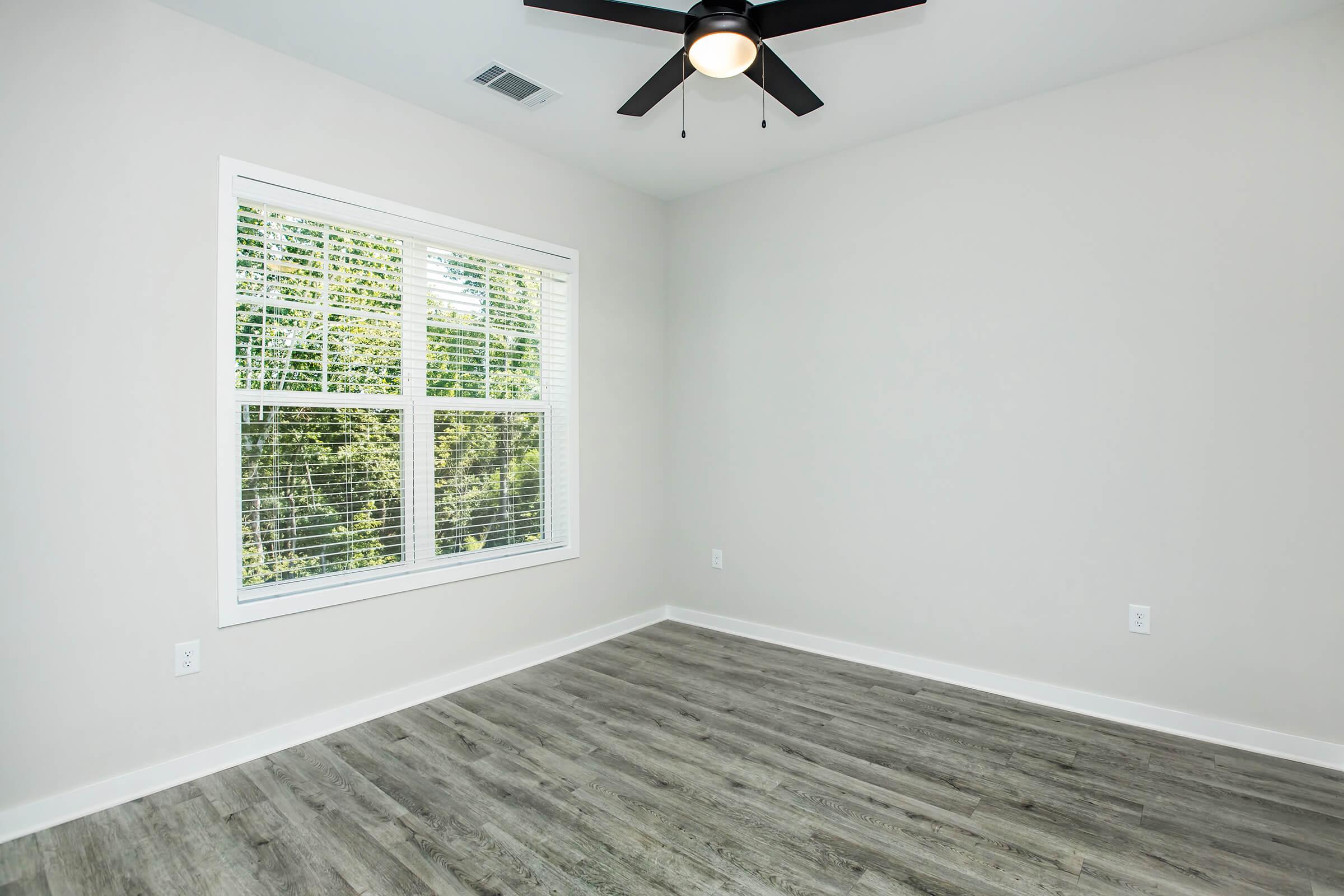 A bright, empty room with light gray walls and a ceiling fan. There is a large window with white blinds, allowing natural light to enter. The flooring is a light wood-like laminate, creating a clean and modern appearance.