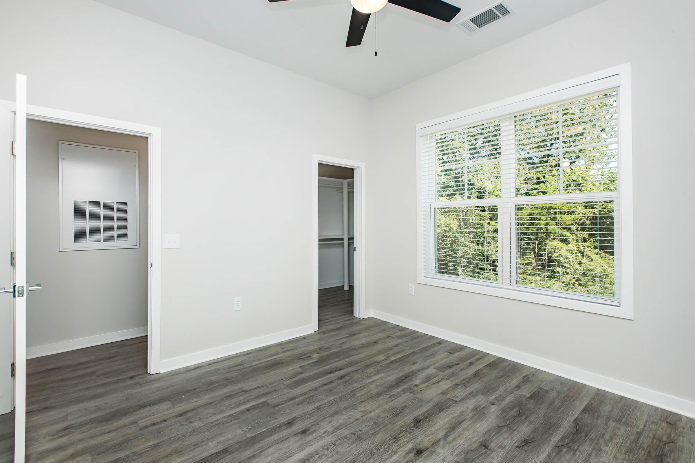 A well-lit, empty room featuring light gray walls, a ceiling fan, and large windows with blinds. The flooring is a laminate wood style, and there's a closet with a door to the left. Another doorway leads to an adjacent space, emphasizing the room's open and spacious feel.