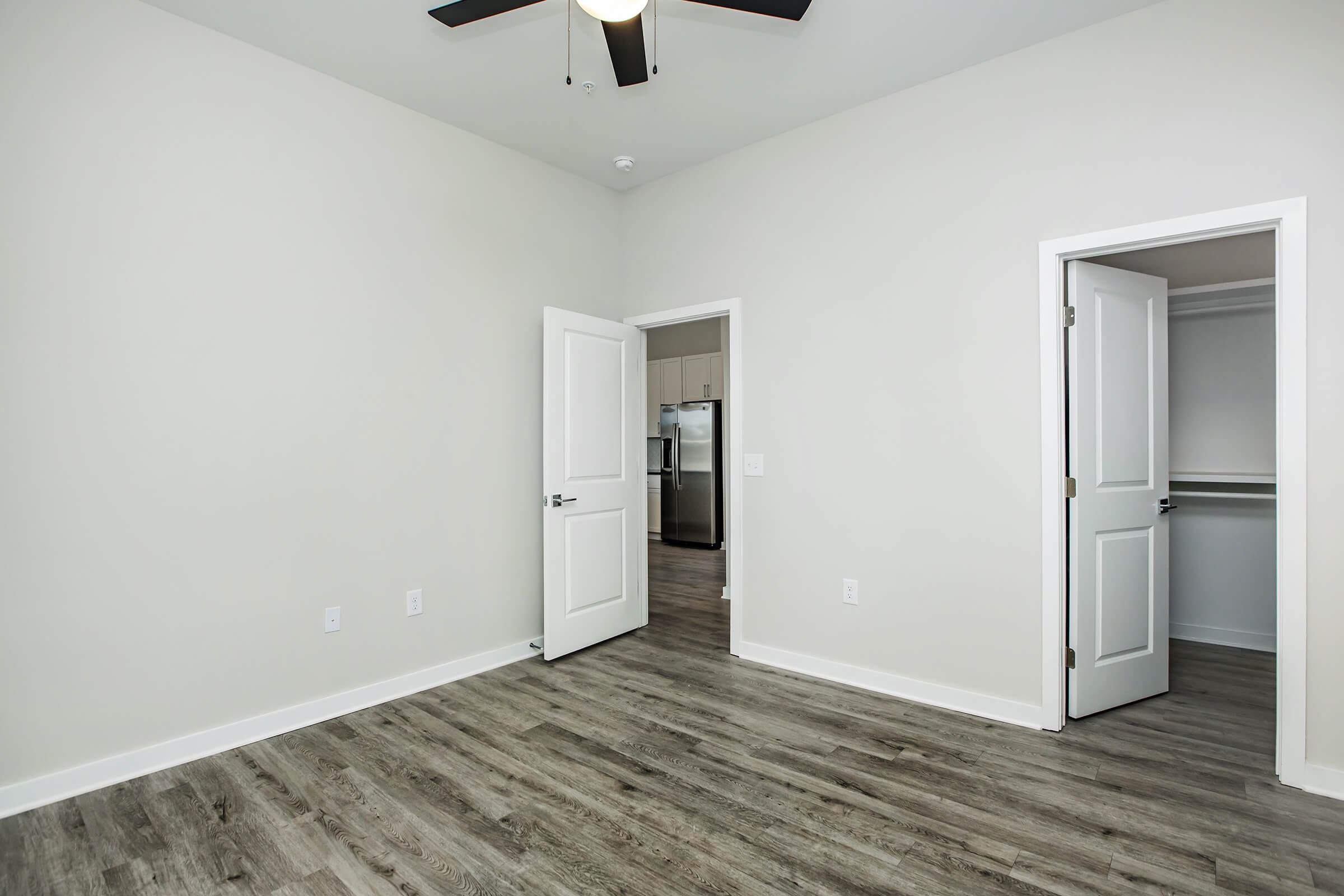 A modern, empty room featuring light-colored walls, a ceiling fan, and laminate flooring. Two open doors lead to a closet and a kitchen area. The space is well-lit, with a neutral color palette, creating a bright and inviting atmosphere suitable for various uses.