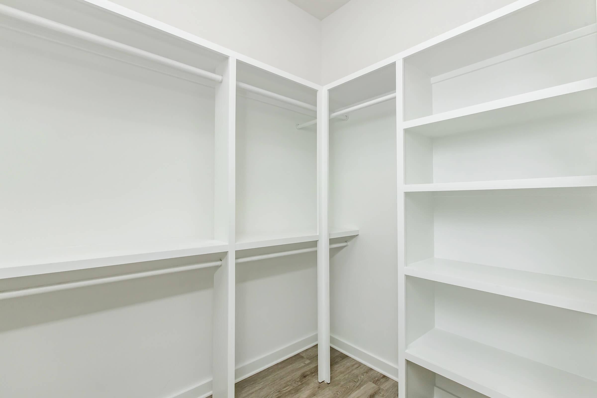 Empty white closet with open shelving and hanging rods, featuring a clean, minimalist design. The walls and shelves are bright and uniform, creating an organized space. The floor has a wood-like finish, adding warmth to the otherwise modern aesthetic.