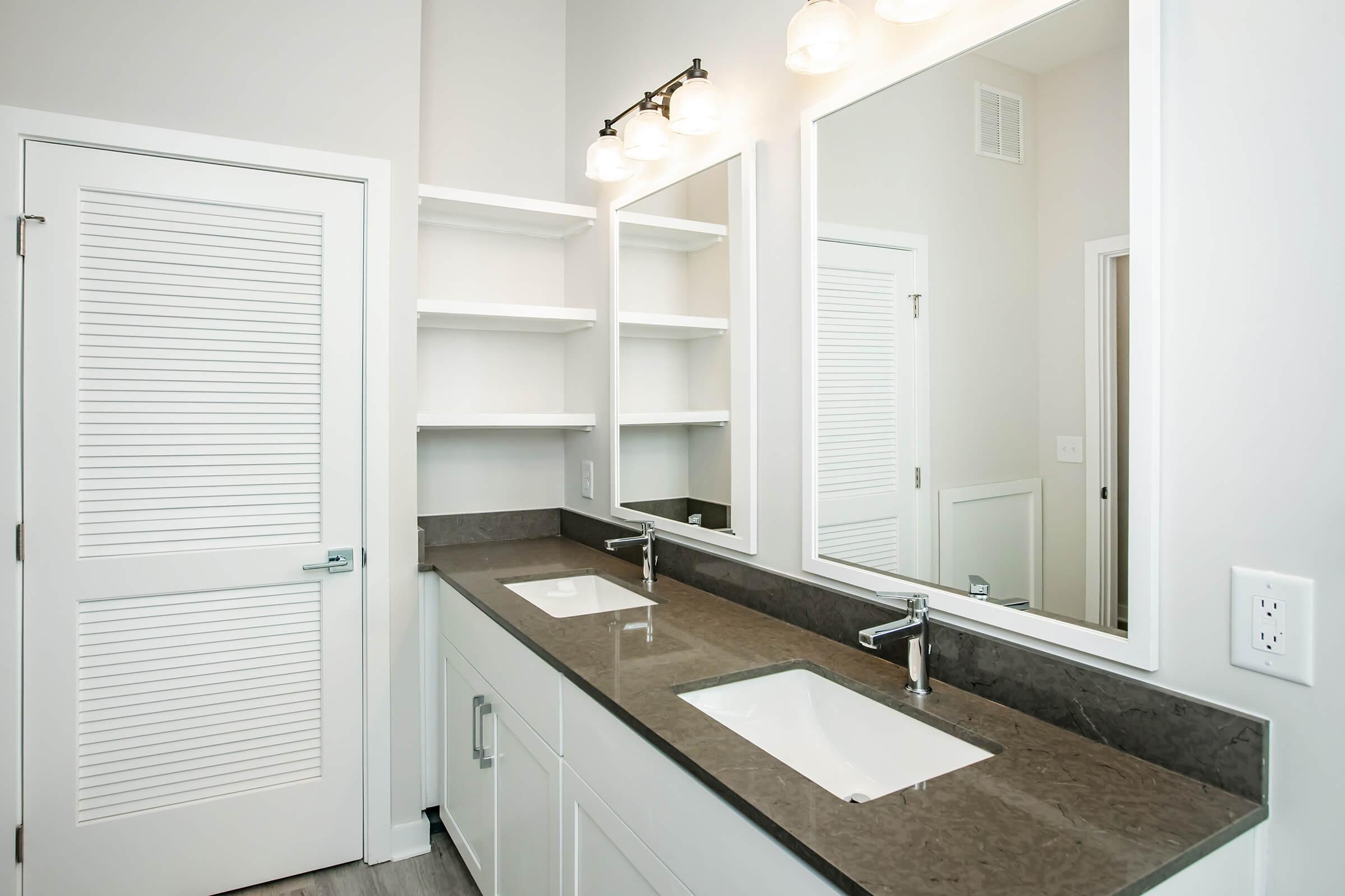 A modern bathroom with a double sink vanity, two mirrors above the sinks, and a light fixture with three bulbs. The countertop is dark and sleek, while the cabinetry is white. There are open shelves to the side and a door with louvered panels leading to another area. The overall design is clean and contemporary.