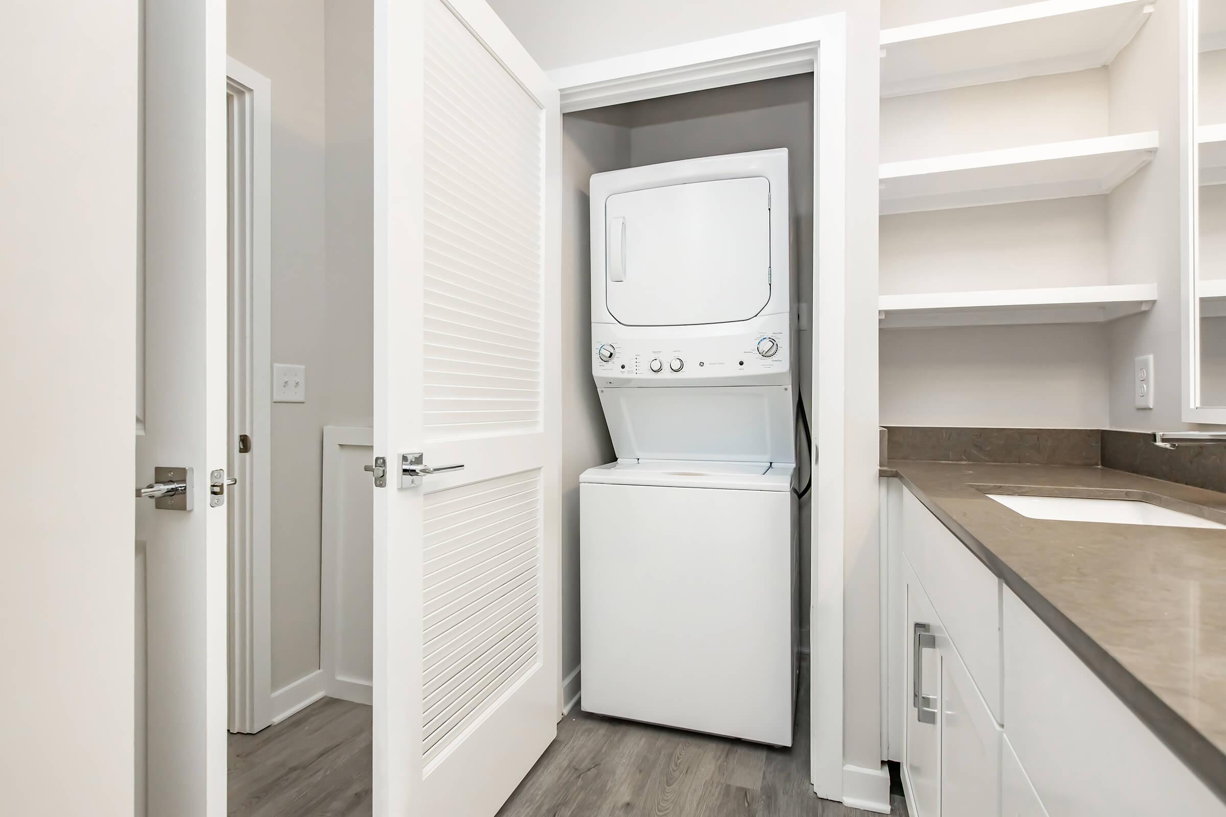 A compact laundry area featuring a stacked washer and dryer in a closet. The space is modern with light-colored walls, a dark countertop, and open shelving. A door partially opened leads to another room, enhancing the home's functional design.