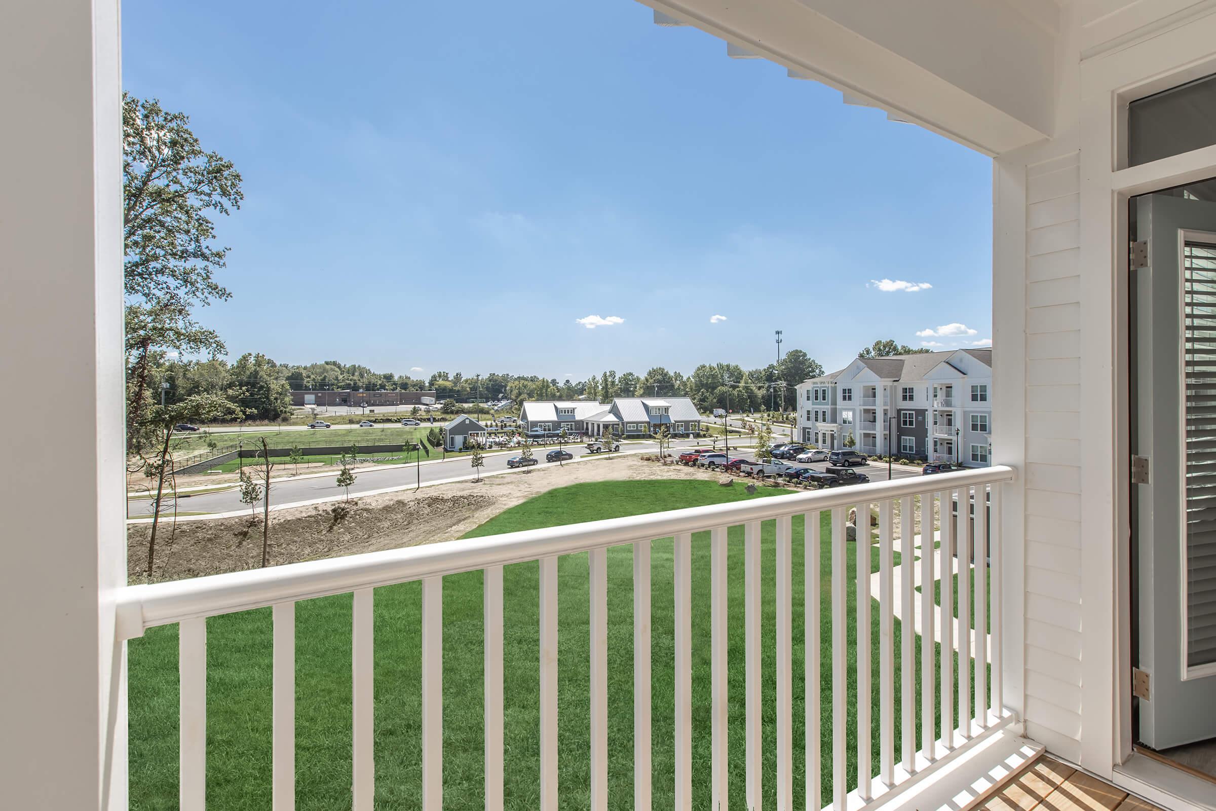 View from a balcony overlooking a landscaped area with green grass, a road, and nearby buildings. The sky is clear and blue, with a few clouds, creating a bright and sunny atmosphere.