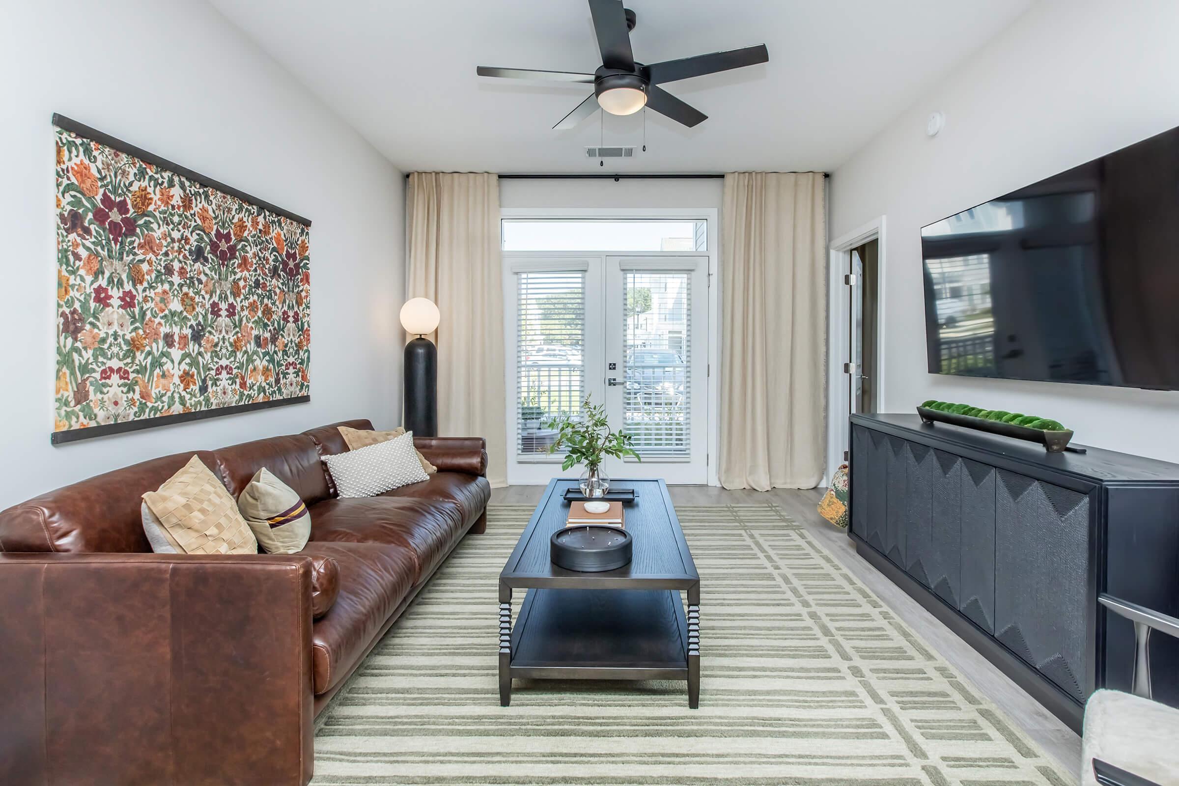 A modern living room featuring a brown leather sofa adorned with decorative pillows, a sleek coffee table, and a large flat-screen TV on a dark cabinet. The walls are painted light gray, and there’s a colorful floral artwork above the sofa. Large windows are covered with light curtains, allowing natural light to enter.
