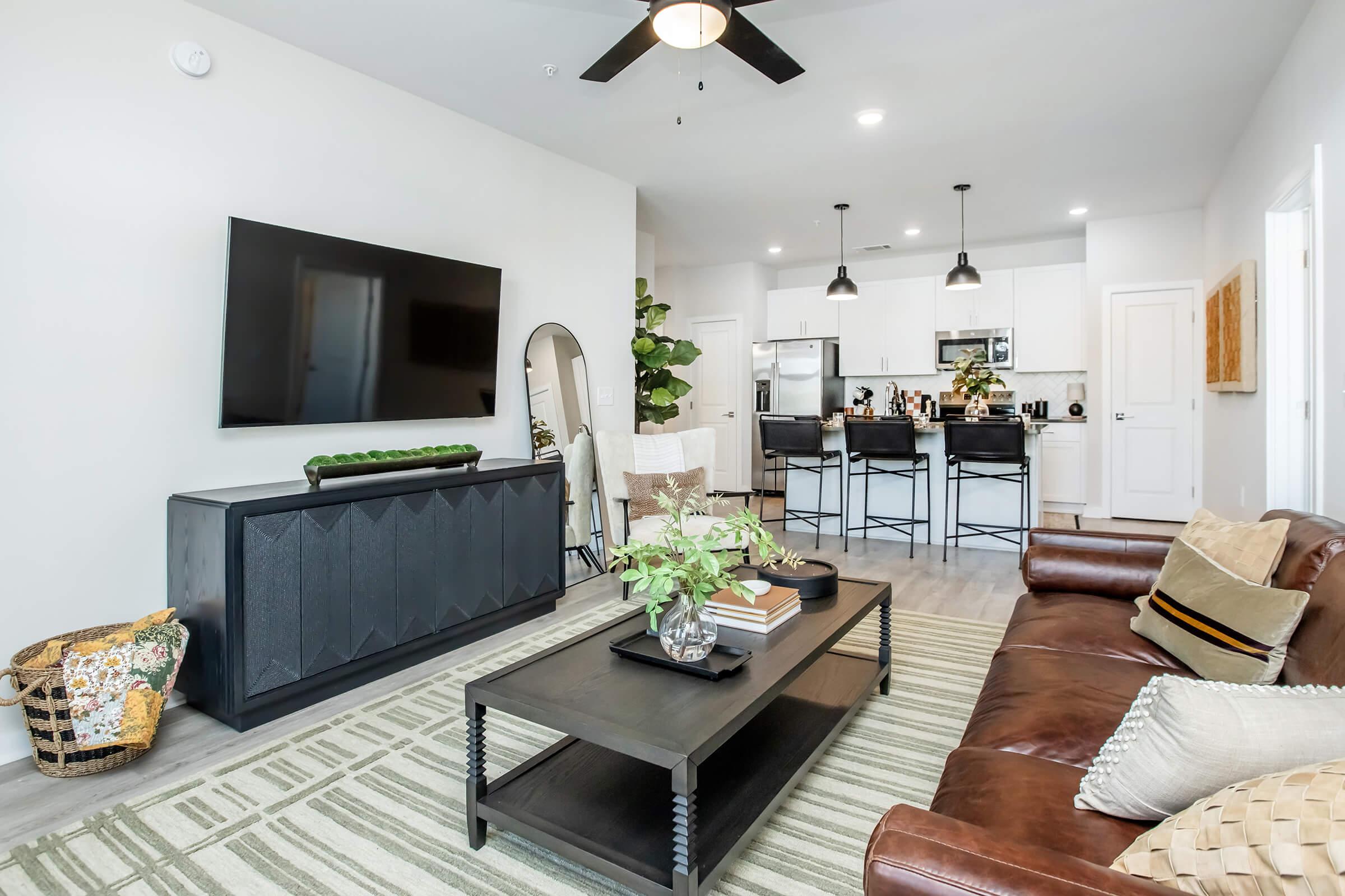 A modern living room featuring a brown leather sofa, a dark wooden coffee table, and a large flat-screen TV on the wall. There is a potted plant and decorative items on the table. The space has a neutral color scheme with a striped area rug and a stylish dining area in the background.