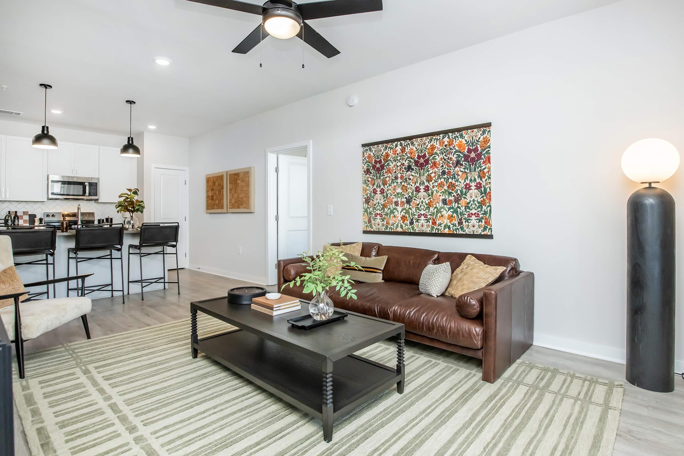 A modern living room with a brown leather sofa adorned with decorative pillows, a coffee table, and a green and white area rug. A colorful wall art piece adds visual interest. There is a tall black lamp and a dining area with black barstools in the background, complemented by a light, airy atmosphere.