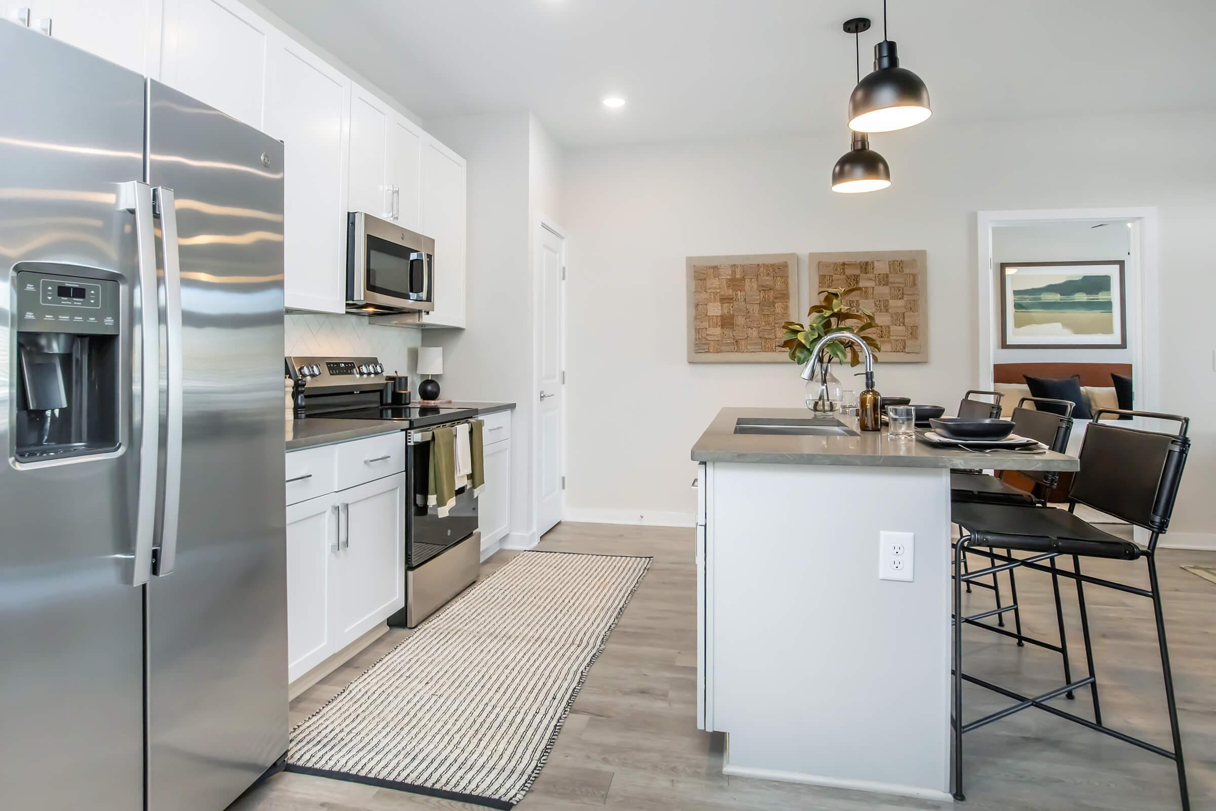 Modern kitchen featuring stainless steel appliances, including a refrigerator and microwave. There is a central island with a countertop, black bar stools, and decorative items. The walls are painted in light colors, and artwork is displayed. A rug is positioned on the floor, enhancing the contemporary feel.