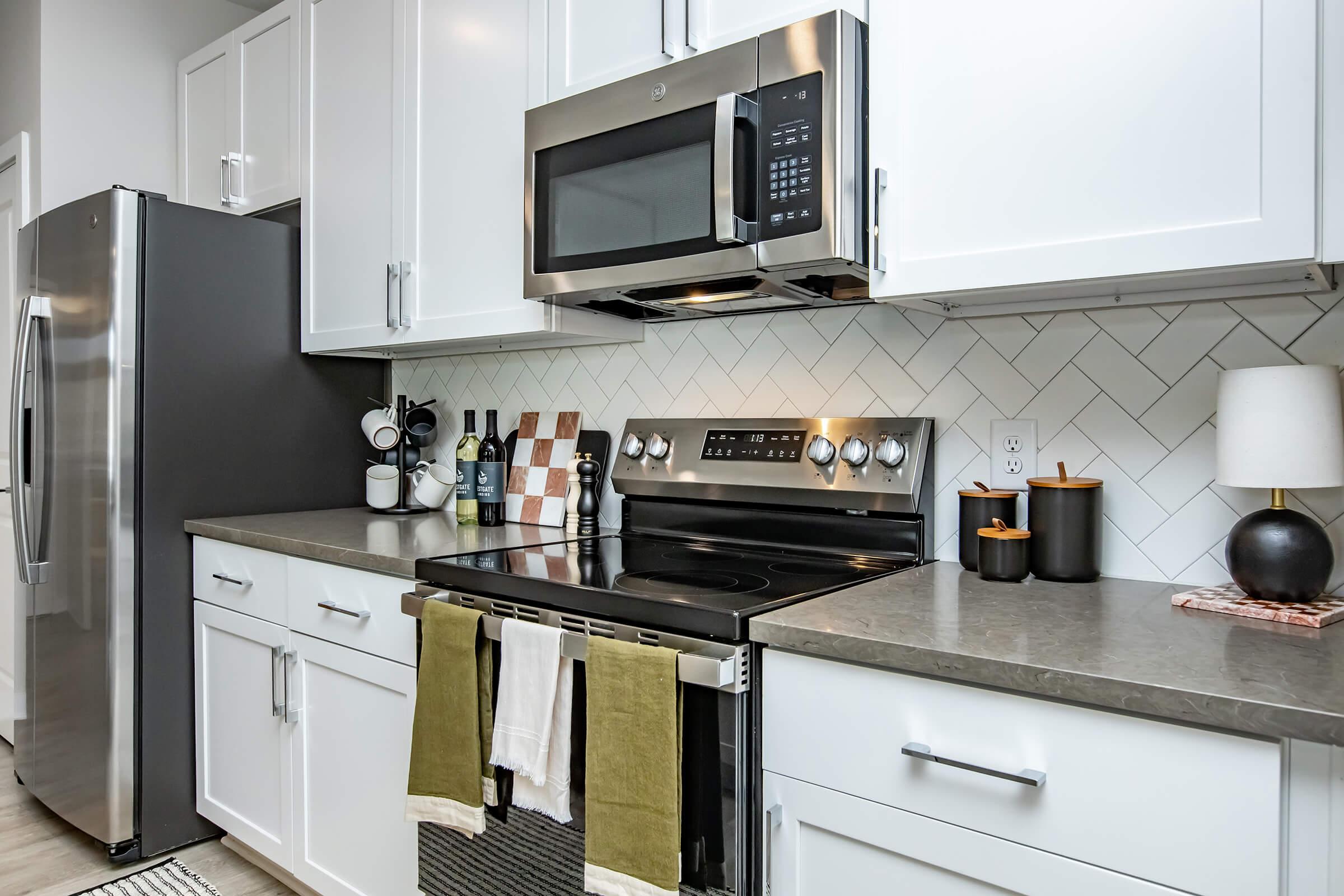 Modern kitchen featuring stainless steel appliances, including a refrigerator and microwave. The countertop is gray with herringbone tile backsplash. A black stove with hanging towels and decorative kitchen items, including bottles and a lamp, create a cozy atmosphere.