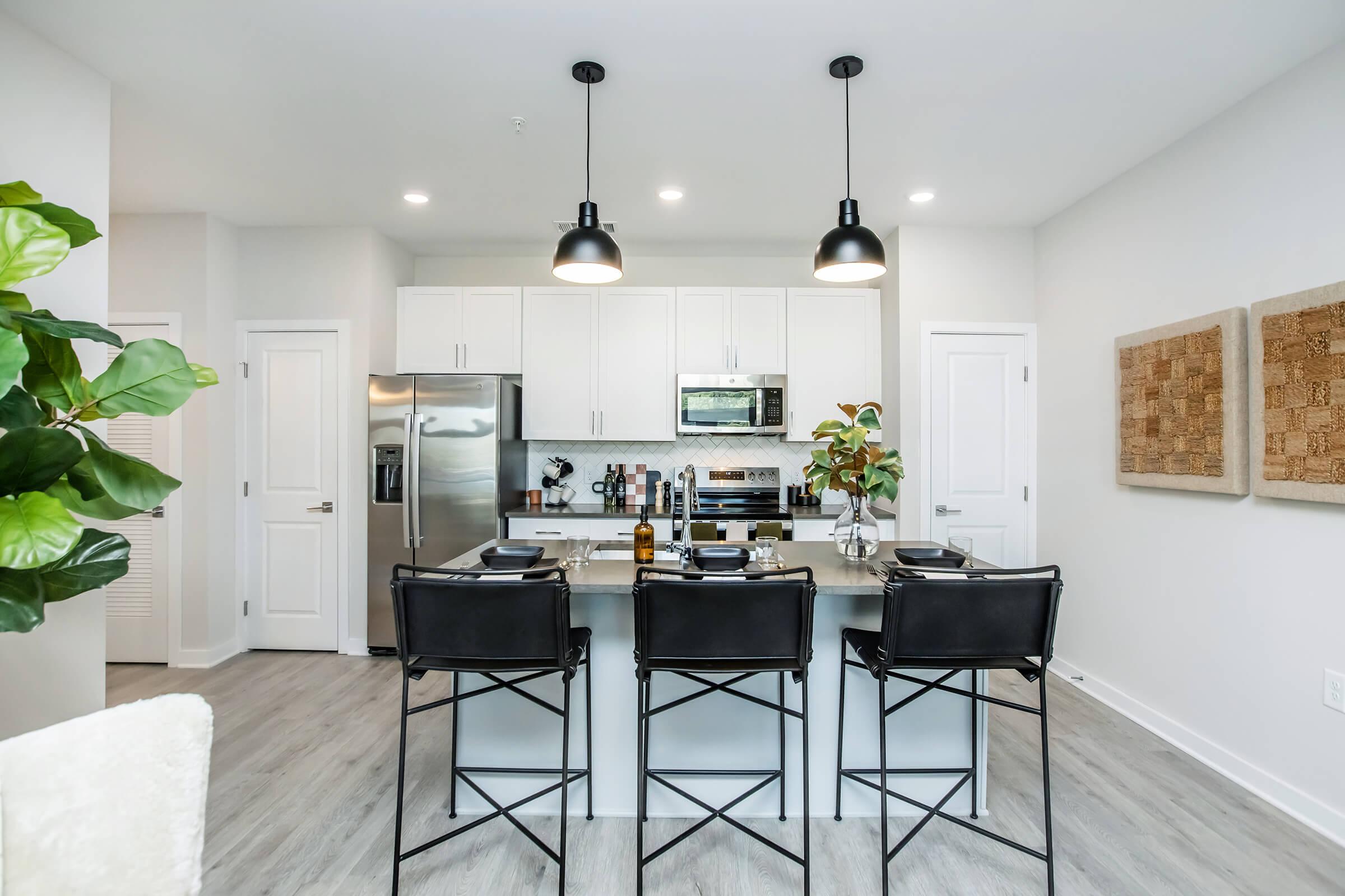 Modern kitchen with white cabinetry, stainless steel appliances, and a large island with four black bar stools. The decor includes a potted plant and wall art made of textured materials. Warm lighting from pendant lights creates a cozy ambiance.