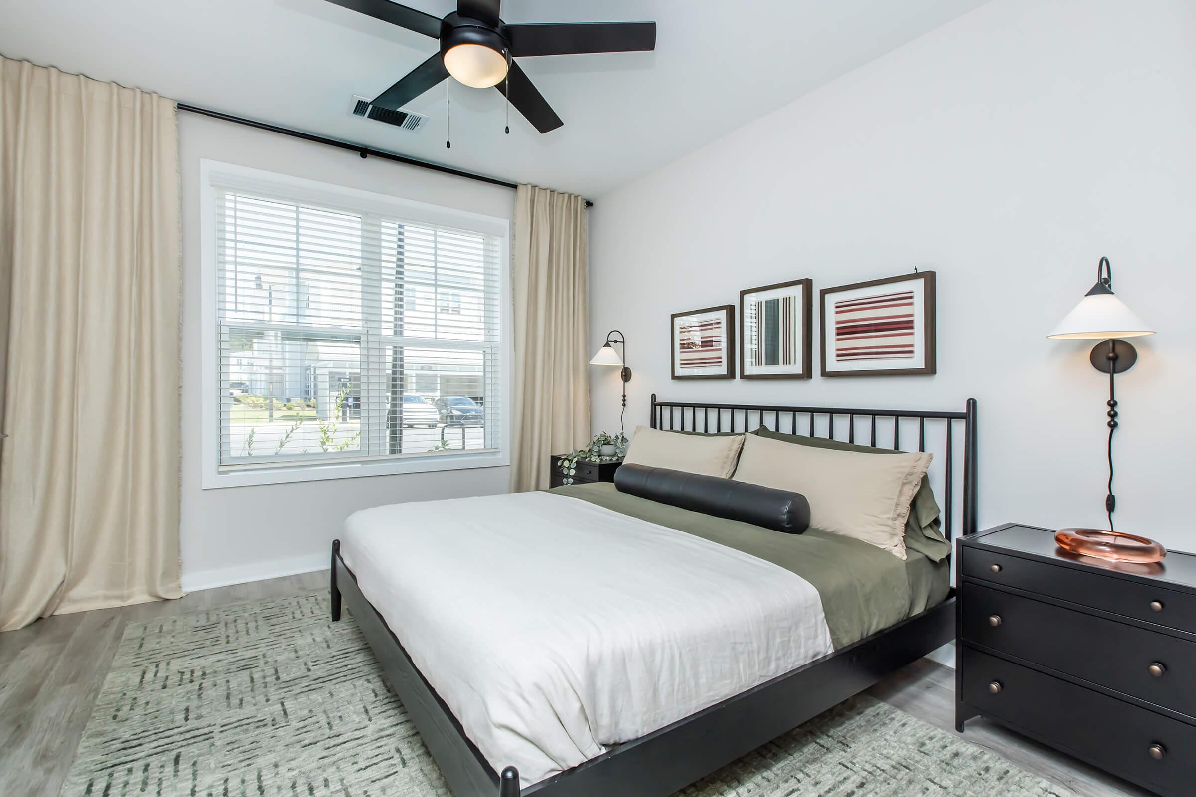 A modern bedroom featuring a black metal bed frame with a white and olive green bedding set. There are two decorative framed prints on the wall, a bedside table with a lamp, and large windows with white blinds allowing natural light in. The floor is covered with a light-colored rug, and soft curtains frame the windows.