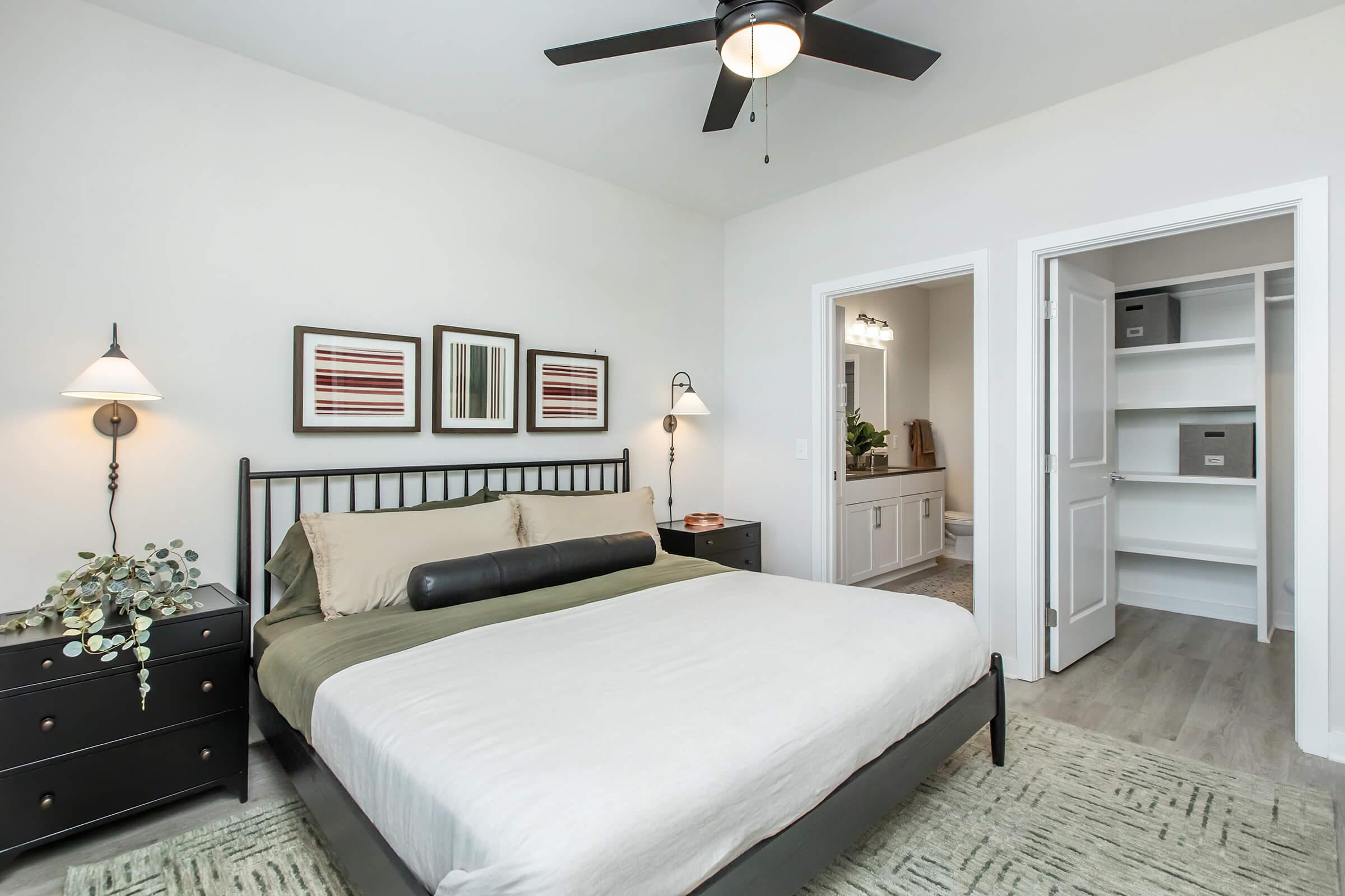 A neatly arranged bedroom featuring a black metal bed frame with a beige and green bedding set. Two framed artworks are on the wall, and the room includes a black nightstand with a plant and lamp. A door leads to a bathroom area, while another door opens to a closet with shelving. The room has a light, modern aesthetic.