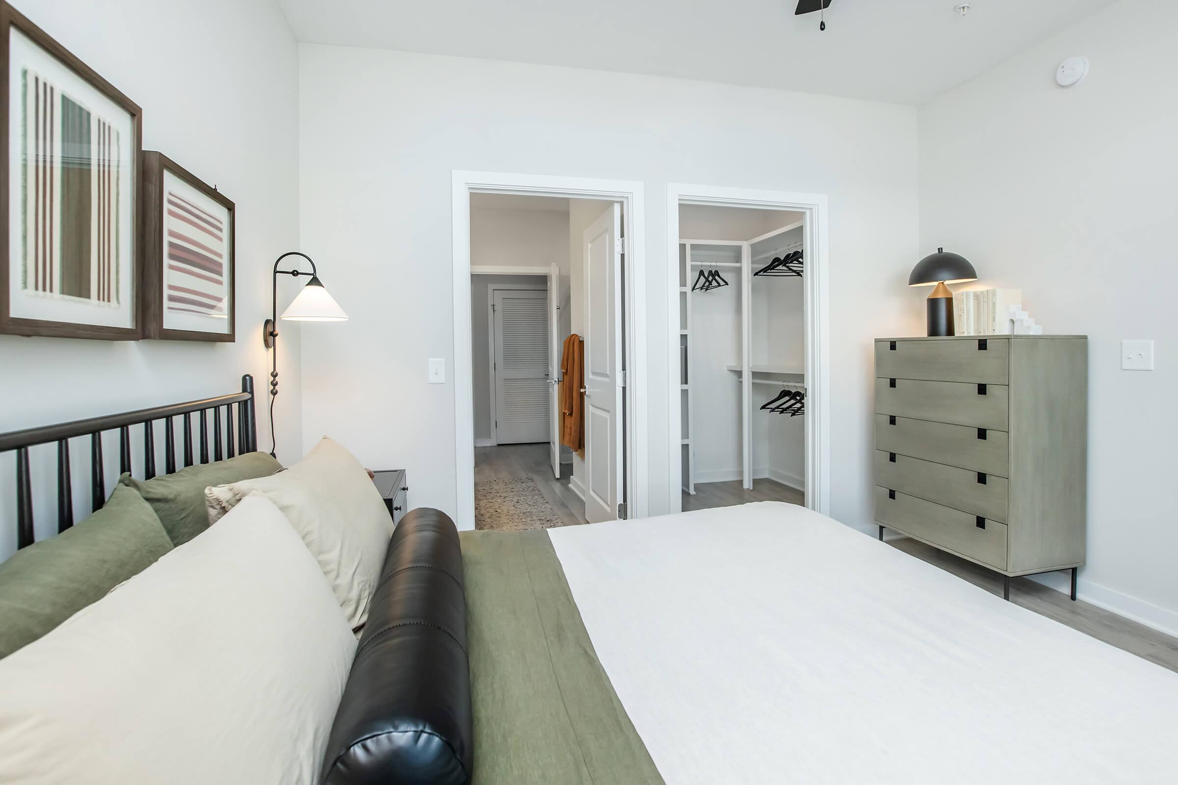 A modern bedroom featuring a black metal bed frame with beige and green bedding. There are framed wall art pieces, a sleek dresser, and a lamp. Two doors lead to closets, and the room has a light, airy feel with neutral walls and a soft rug on the floor.