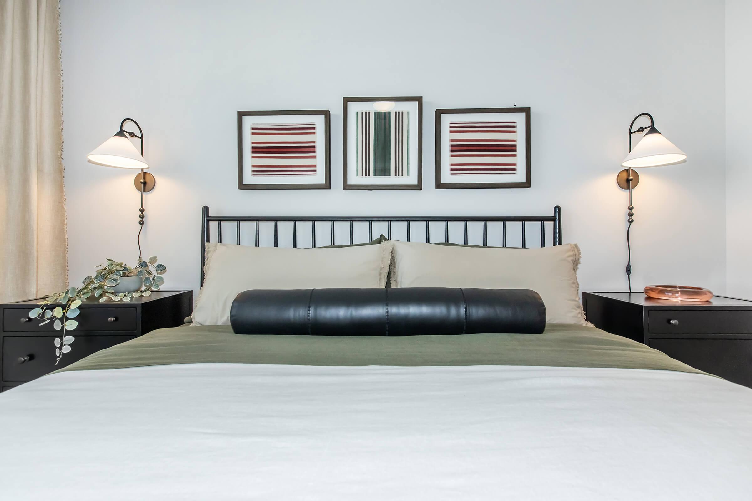 A cozy bedroom featuring a black metal bed frame with two beige pillows and a black bolster pillow. On either side of the bed are dark nightstands with lamps. Above the bed are three framed artworks with abstract striped designs. A green throw is casually draped over the bed, adding a touch of color to the serene space.