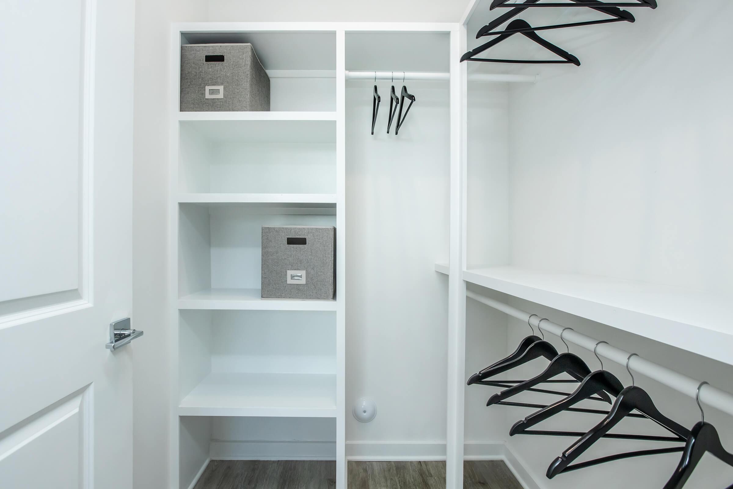 A clean and organized closet featuring white shelves and a hanging rod. One side has several empty black hangers, while the other has two gray storage boxes with labels. The floor is light-colored, complementing the overall minimalist design.