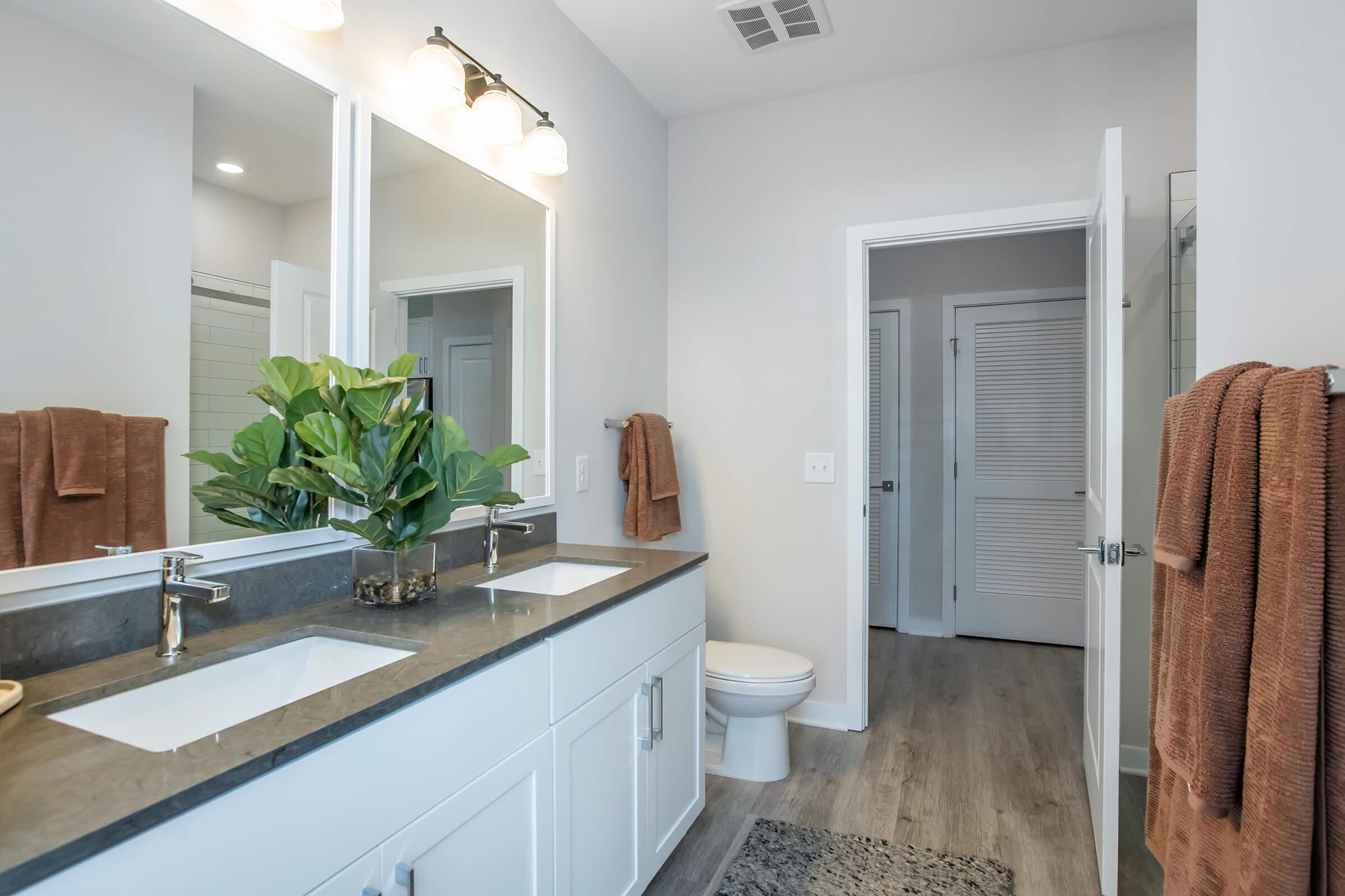 A modern bathroom featuring a double vanity with two sink basins, stylish lighting above, and light gray walls. Towels are neatly hung, and a small plant adds a touch of greenery. A shower area is visible to the left, and a door leads to another room at the back. The flooring is a light wood laminate.
