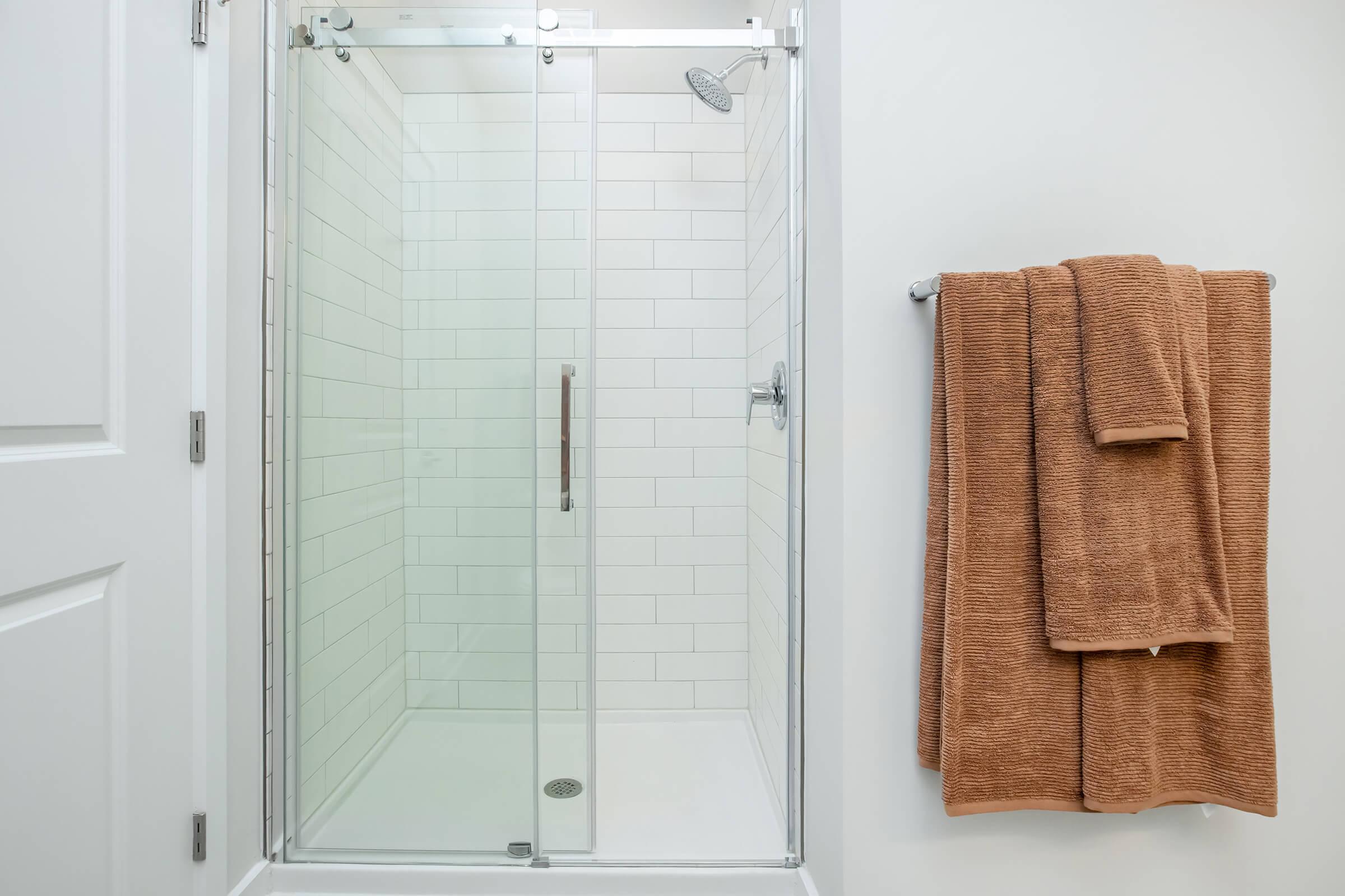 A modern shower with glass sliding doors and white tiled walls. A bronze towel rack holds a plush, rust-colored towel hanging beside the shower. The space is clean and well-lit, featuring a minimalist design.