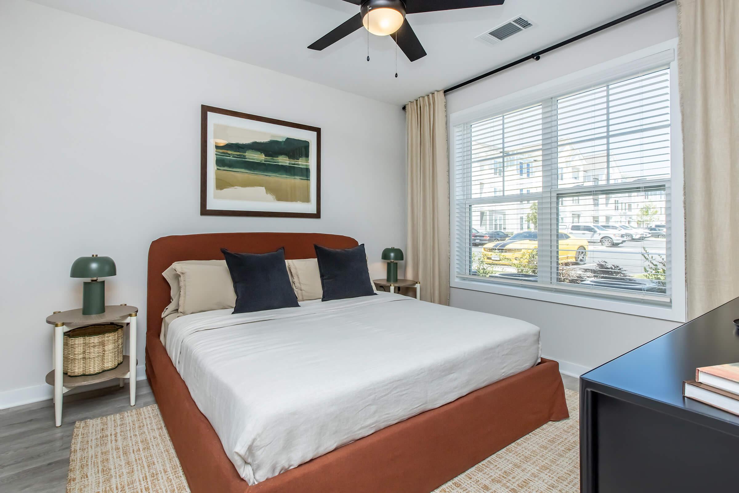A modern bedroom featuring a queen-sized bed with a rust-colored bed frame, two beige pillows, and decorative navy blue cushions. There is a green lamp on a nightstand, a large window with white blinds, and light-colored curtains. A piece of artwork hangs above the bed, and the room has a light rug and a black nightstand.